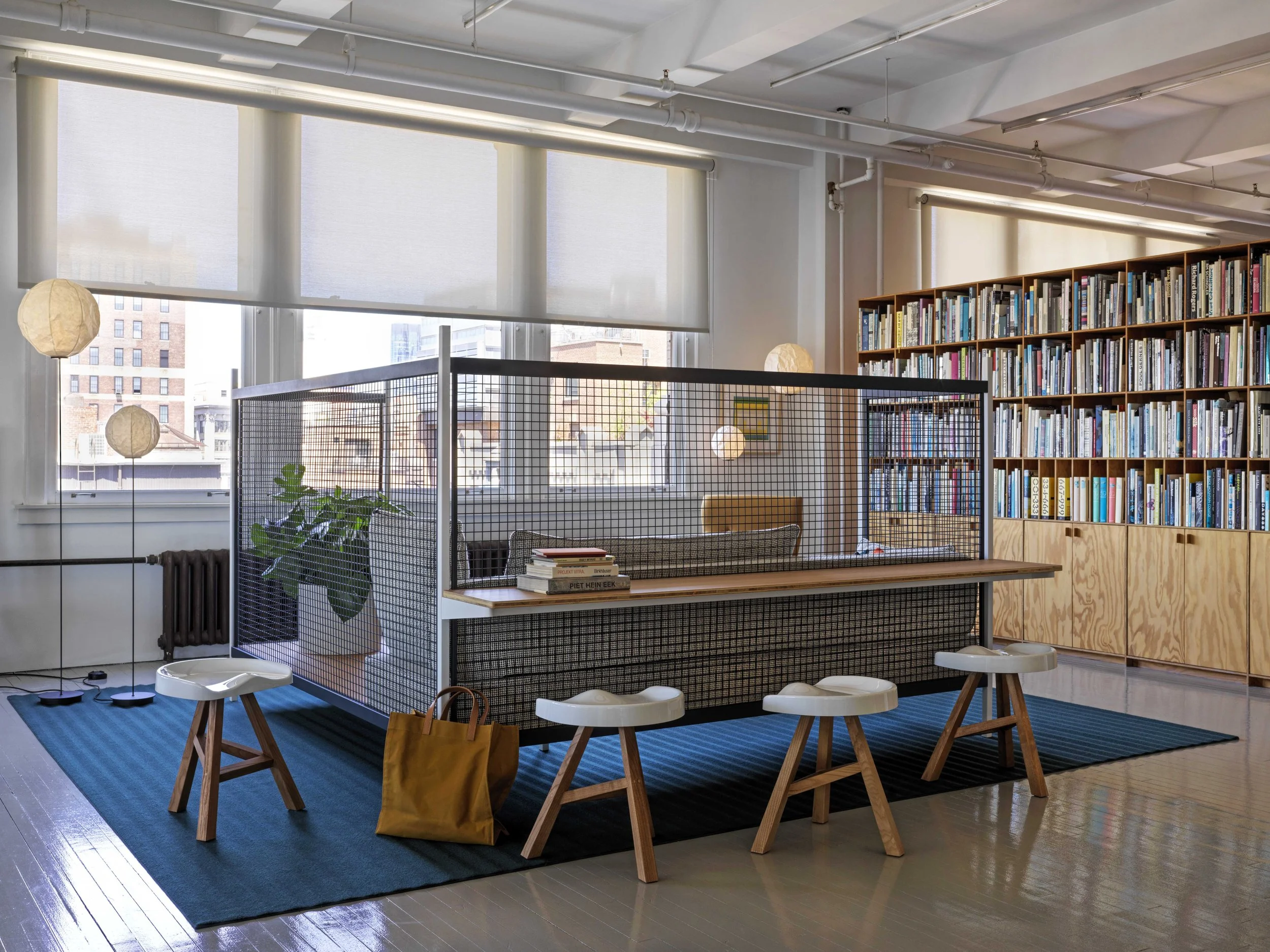 Modern reading nook inside a well-lit room with large windows, a bookcase filled with books, a small table with books, stools, and soft lighting.