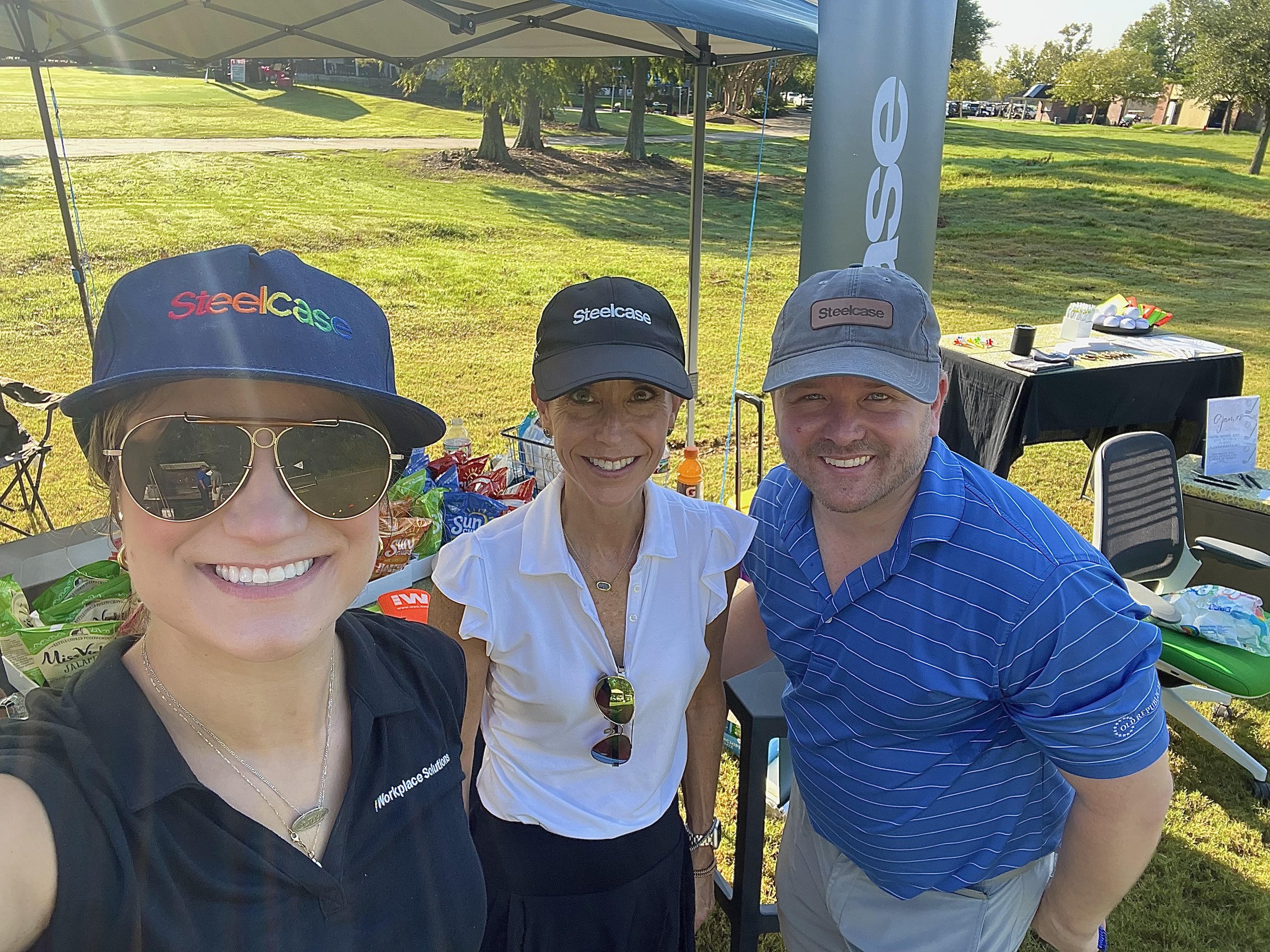 The Workplace Solutions and Steelcase team wearing Steelcase logo hats, taking a selfie under a canopy with snack tables at a corporate golf event.