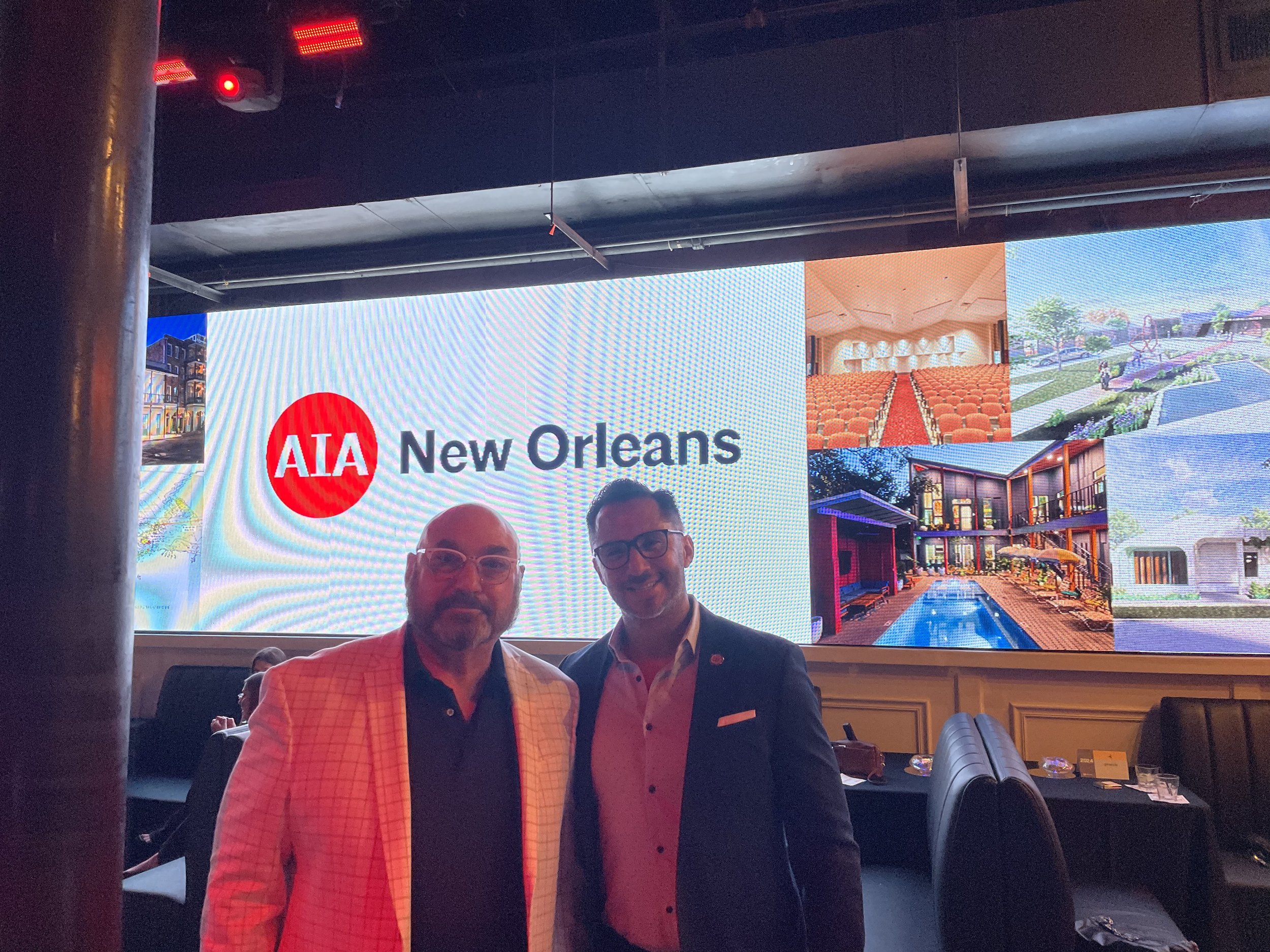 Paul Maczka and Francisco Morales from Workplace Solutions posing for a picture in front of a large screen displaying the AIA New Orleans logo and images at the 2024 AIA Awards event.