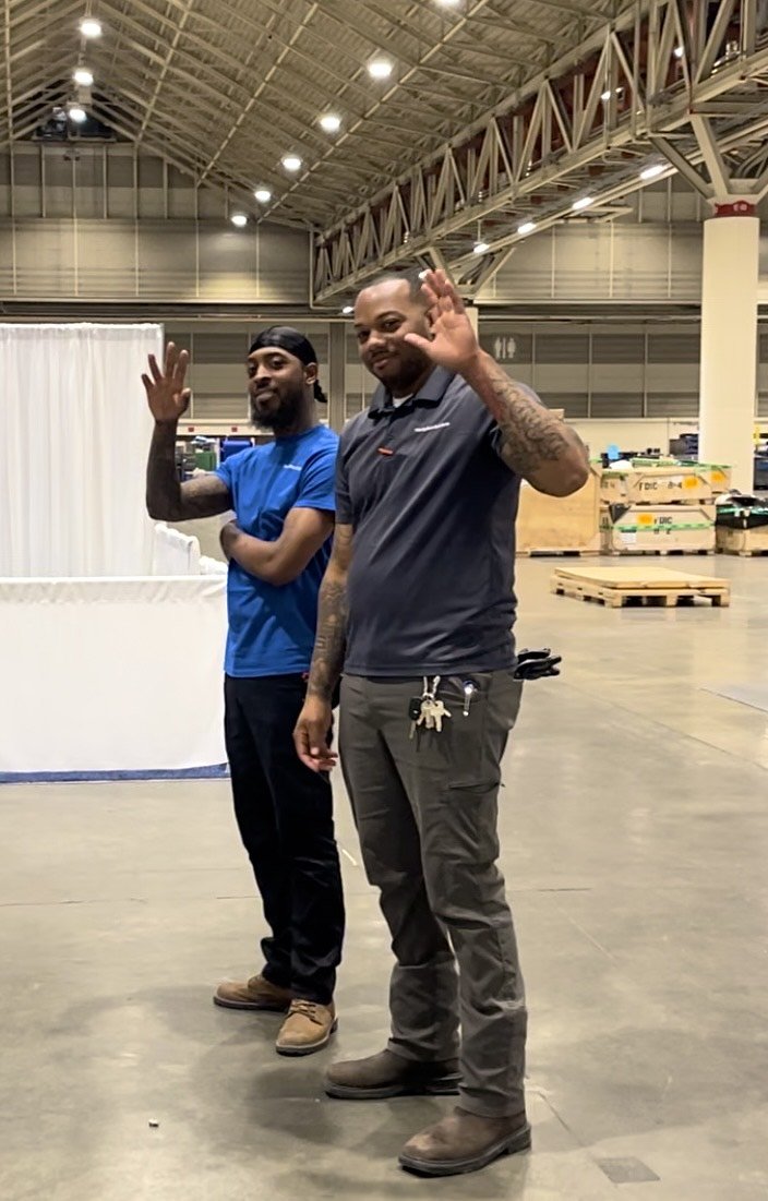 Two members of the New Orleans Workplace Solutions installation team standing inside a large warehouse, waving and smiling at the camera.