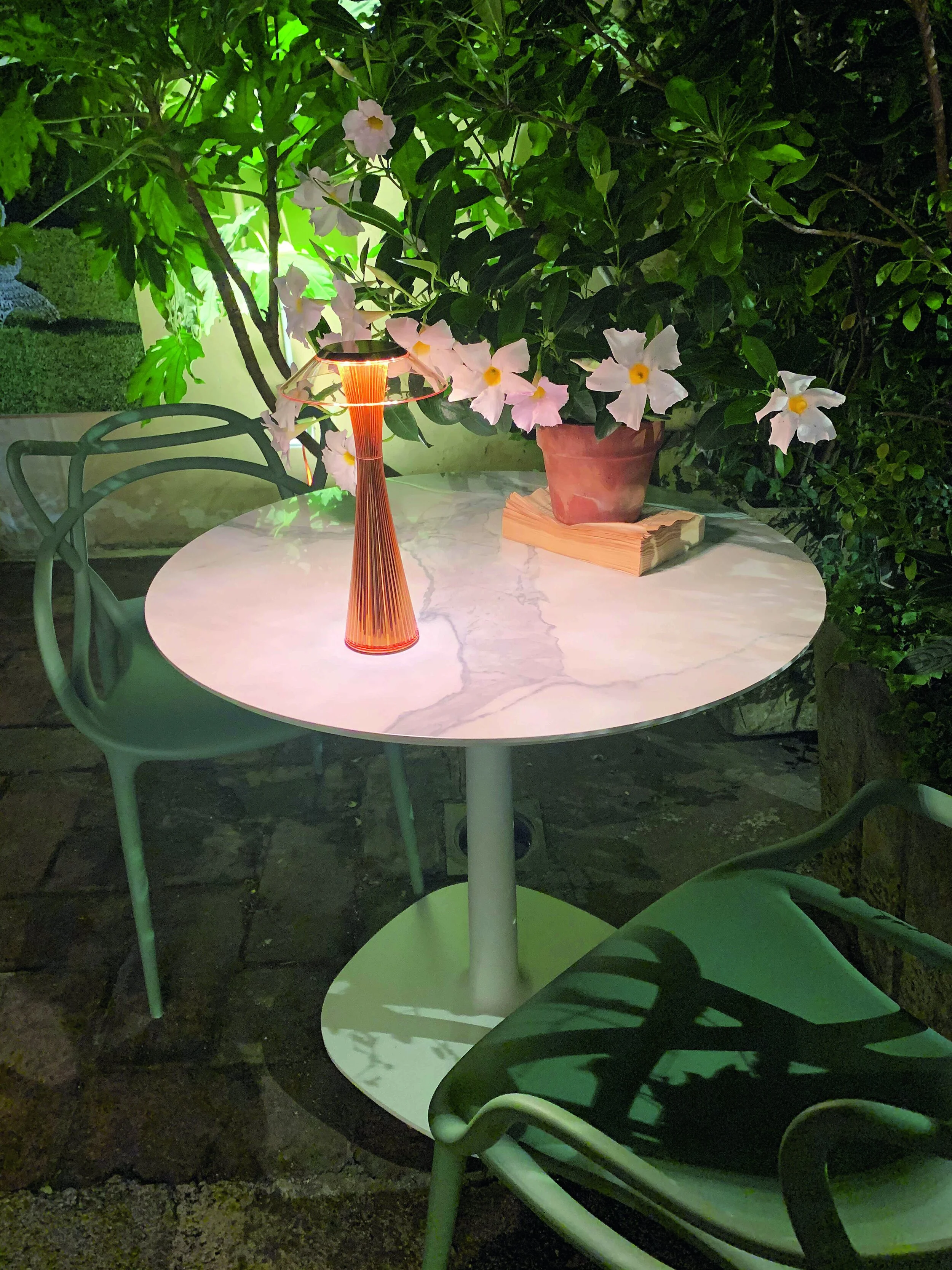 Round white marble Kartell table outdoors with a small red and orange decorative lamp, surrounded by green Kartell chairs.