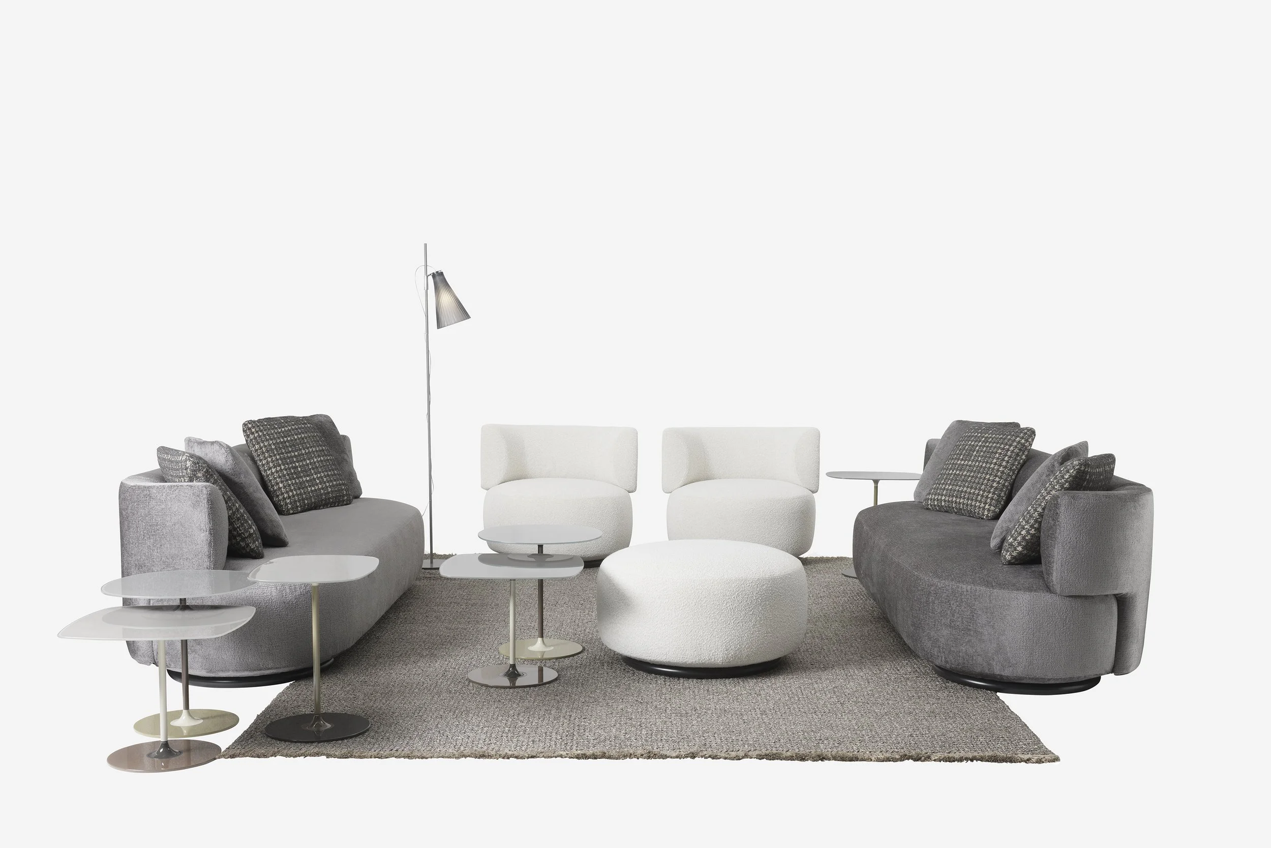 Modern living room with two gray sofas, two white armchairs, a white ottoman, several small side tables, a floor lamp, and a beige rug.