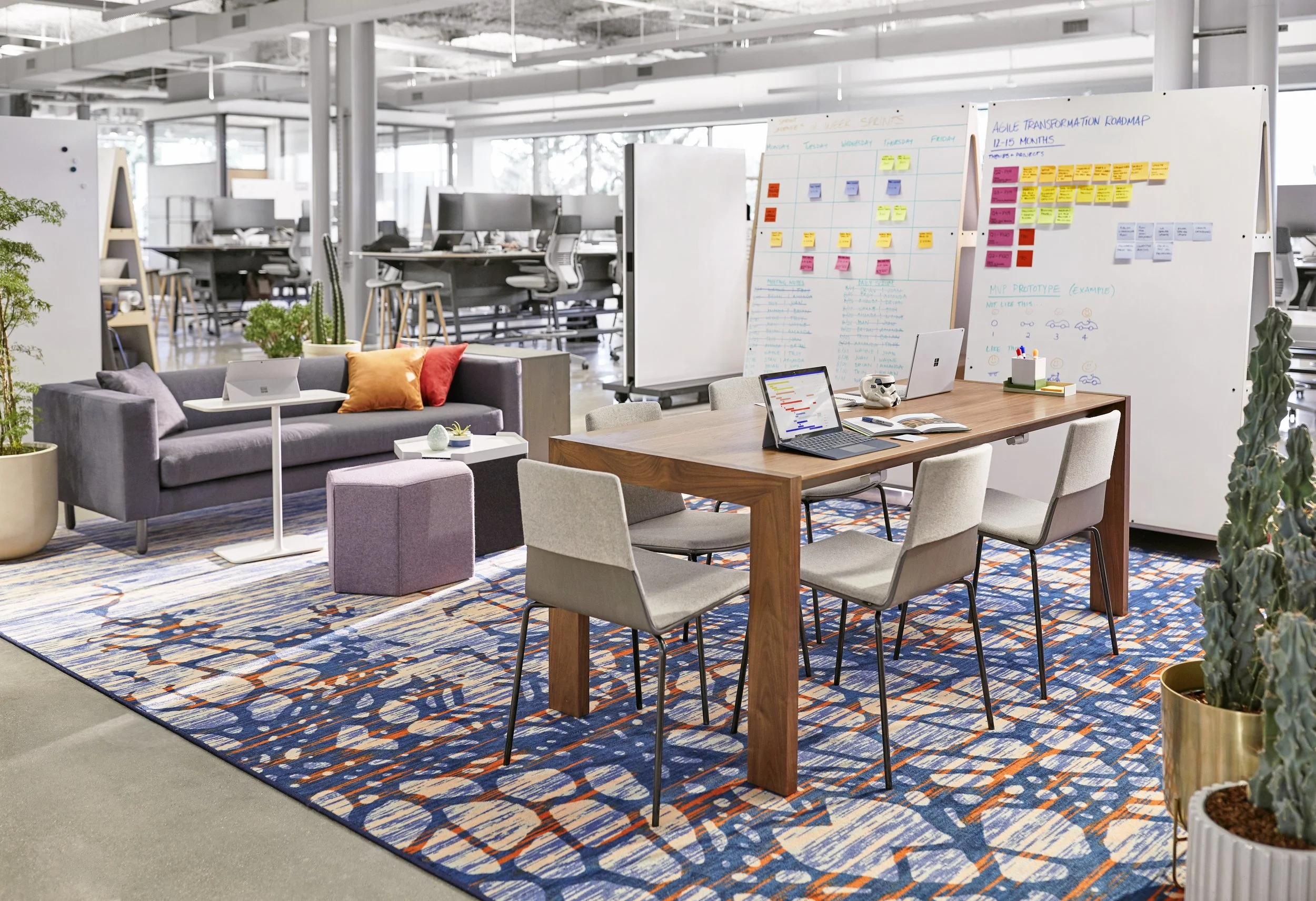 Open office space with a meeting area, including a wooden table with chairs from Blu Dot, laptops, and whiteboards with colorful sticky notes and charts. There is a gray sofa with pillows, plants, and a patterned rug.