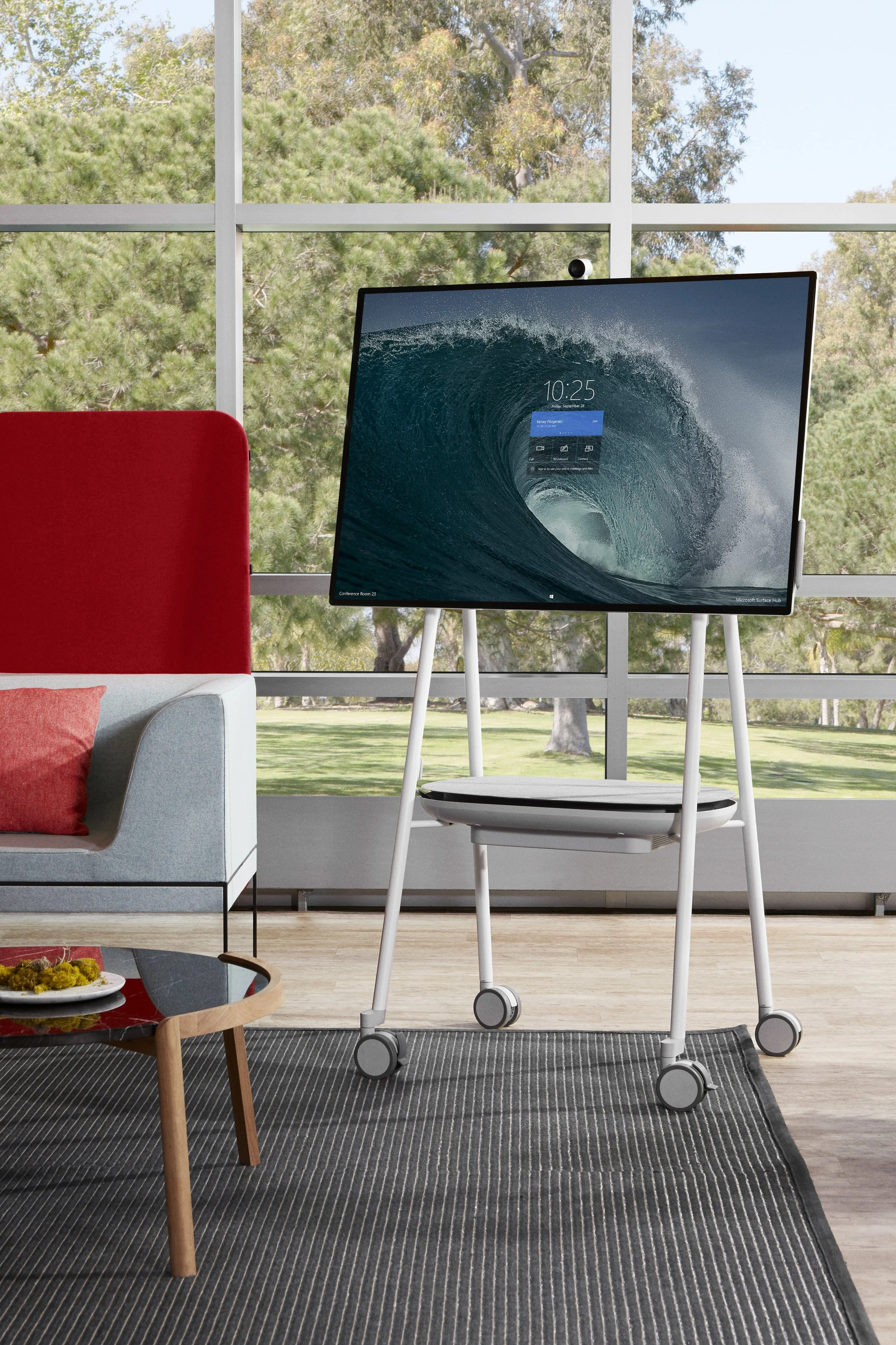 Modern open-office, featuring a flat-screen monitor on a white mobile stand, a light gray sofa with a red cushion, and a wooden coffee table.