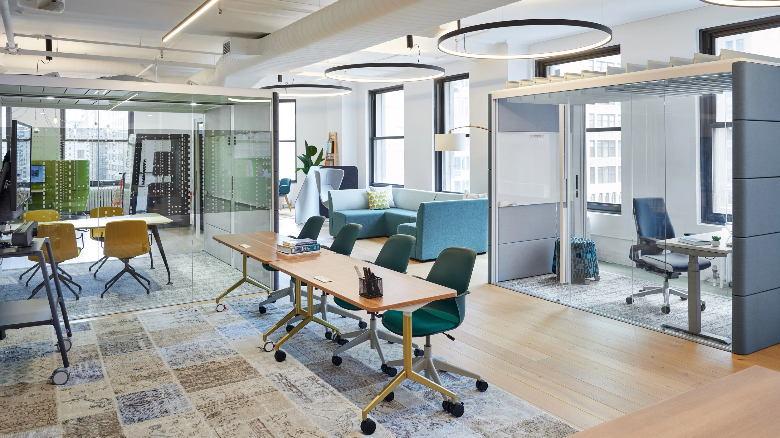 Office featuring Orangebox Air3 private office pod, a wooden conference table with green office chairs nearby, and a seating area with a light blue couch and armchair.