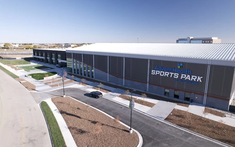 An aerial view of AdvenHealth Sports Park at Blue Hawk, a modern sports facility with a large building, parking lot, and surrounding landscaped areas.