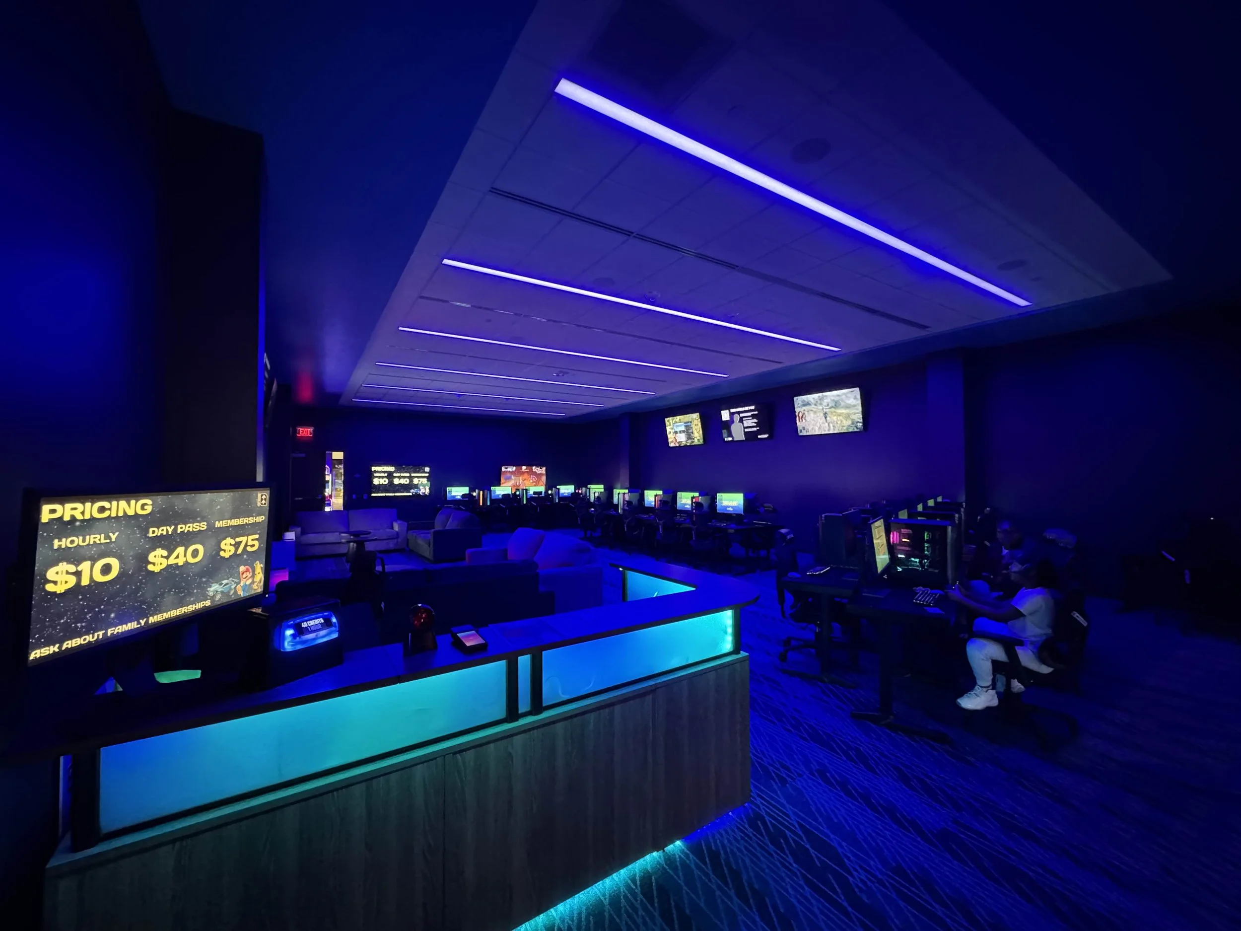 A dimly lit indoor arcade or gaming center with neon blue lighting. There are multiple gaming stations with screens on the right side, some people sitting and playing. The front counter displays pricing information for hourly, day pass, and membership options.