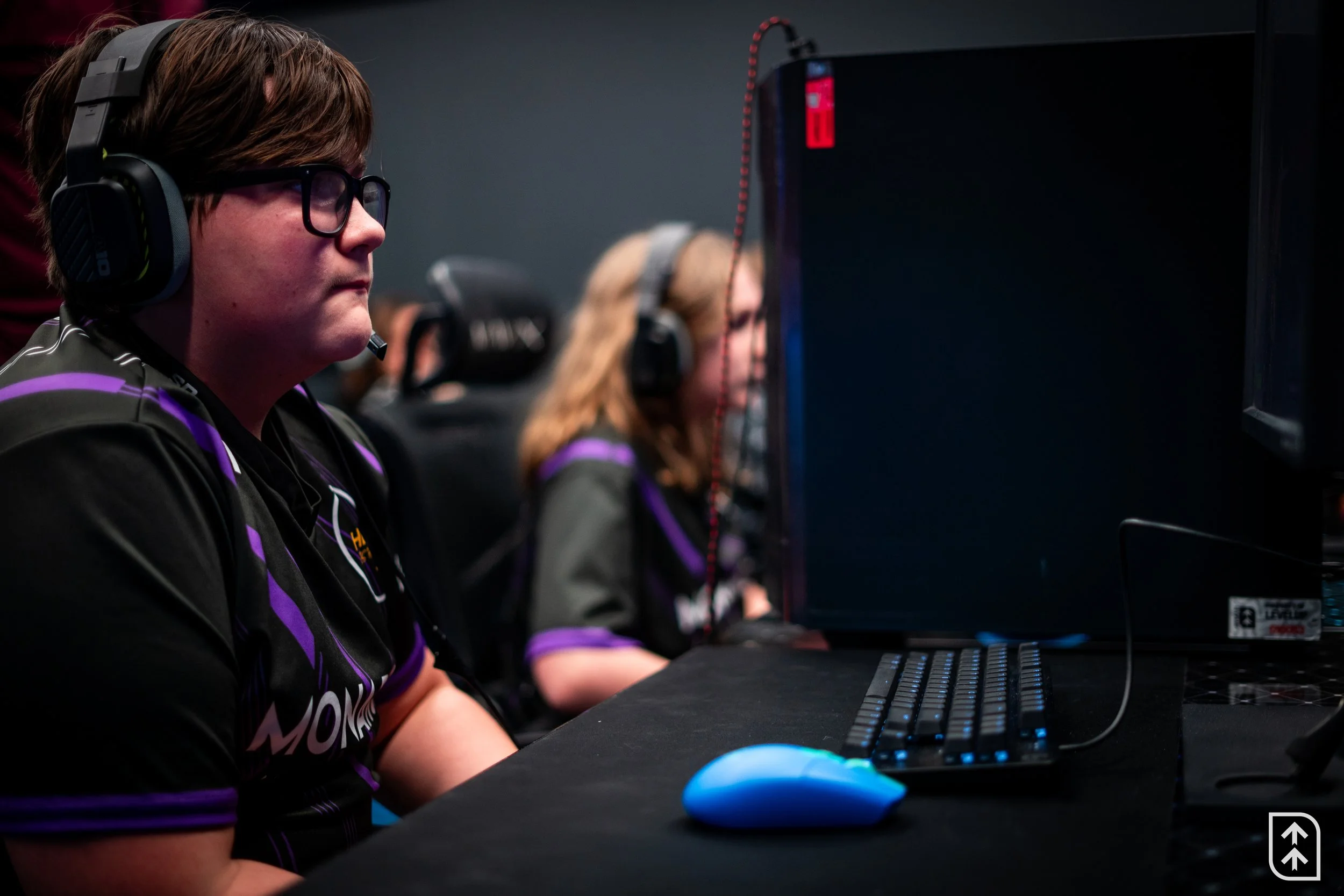 Esports players sitting at computers wearing gaming headsets, focused on screen, in a dark gaming room.