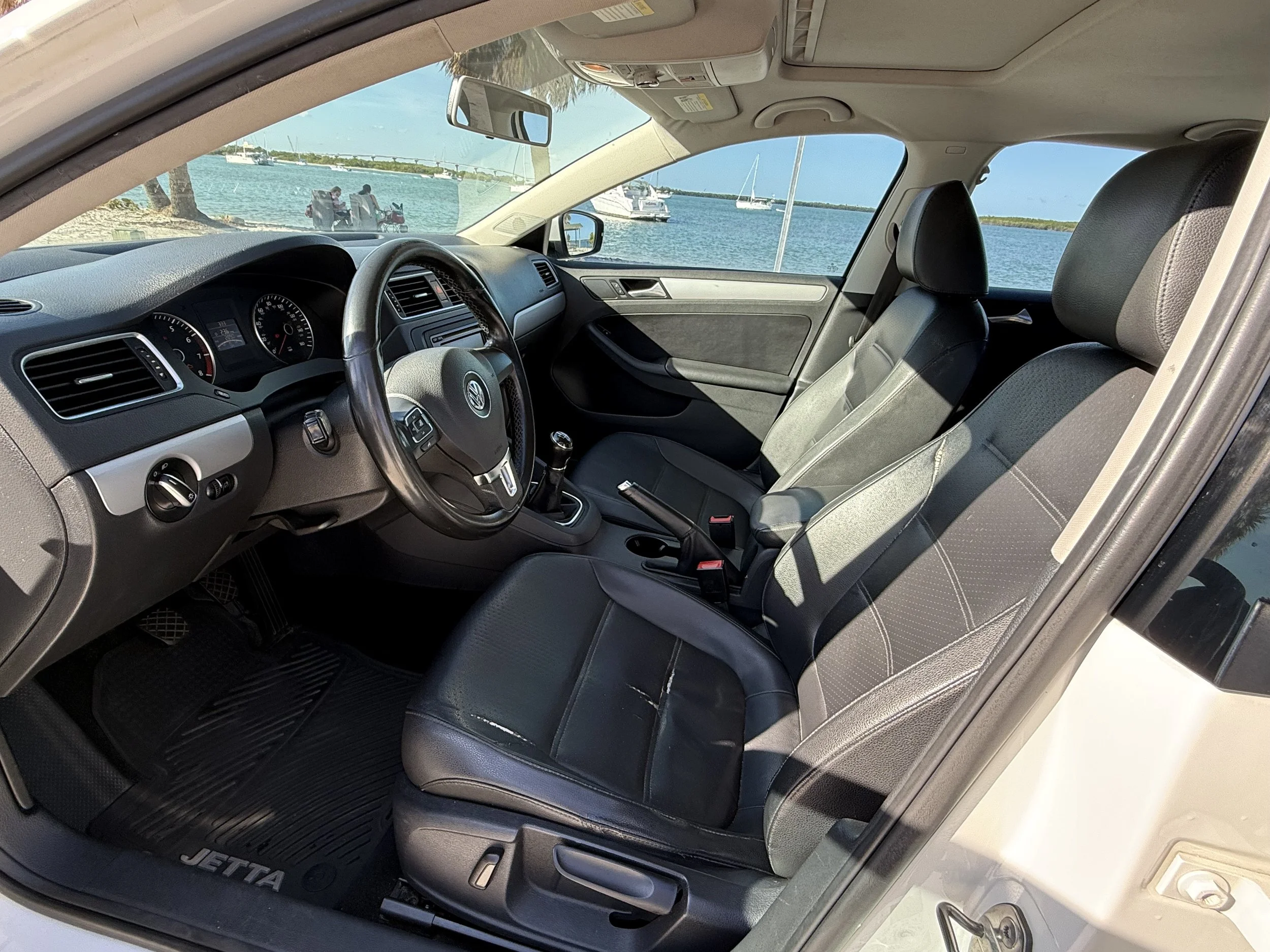 The interior of a Volkswagen Jetta car with black seats, dashboard, and steering wheel, parked near a shoreline with boats and a clear sky visible through the windows.