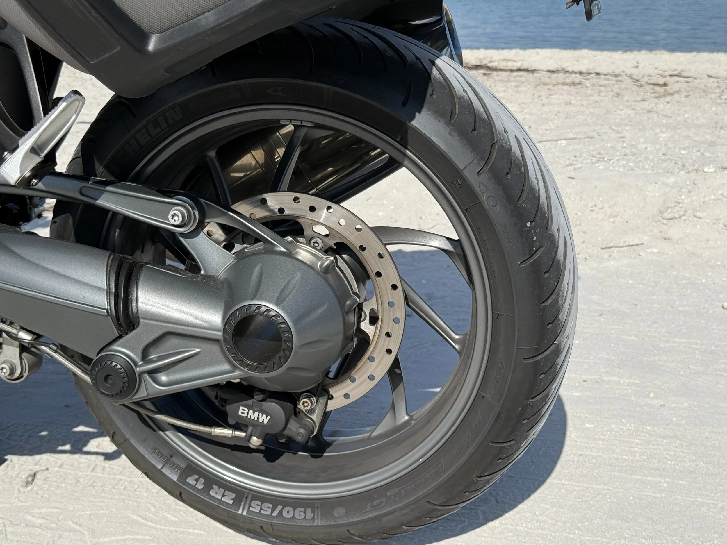 Close-up of a motorcycle rear wheel, tire, disc brake, and part of the chain drive on a sandy surface near water.
