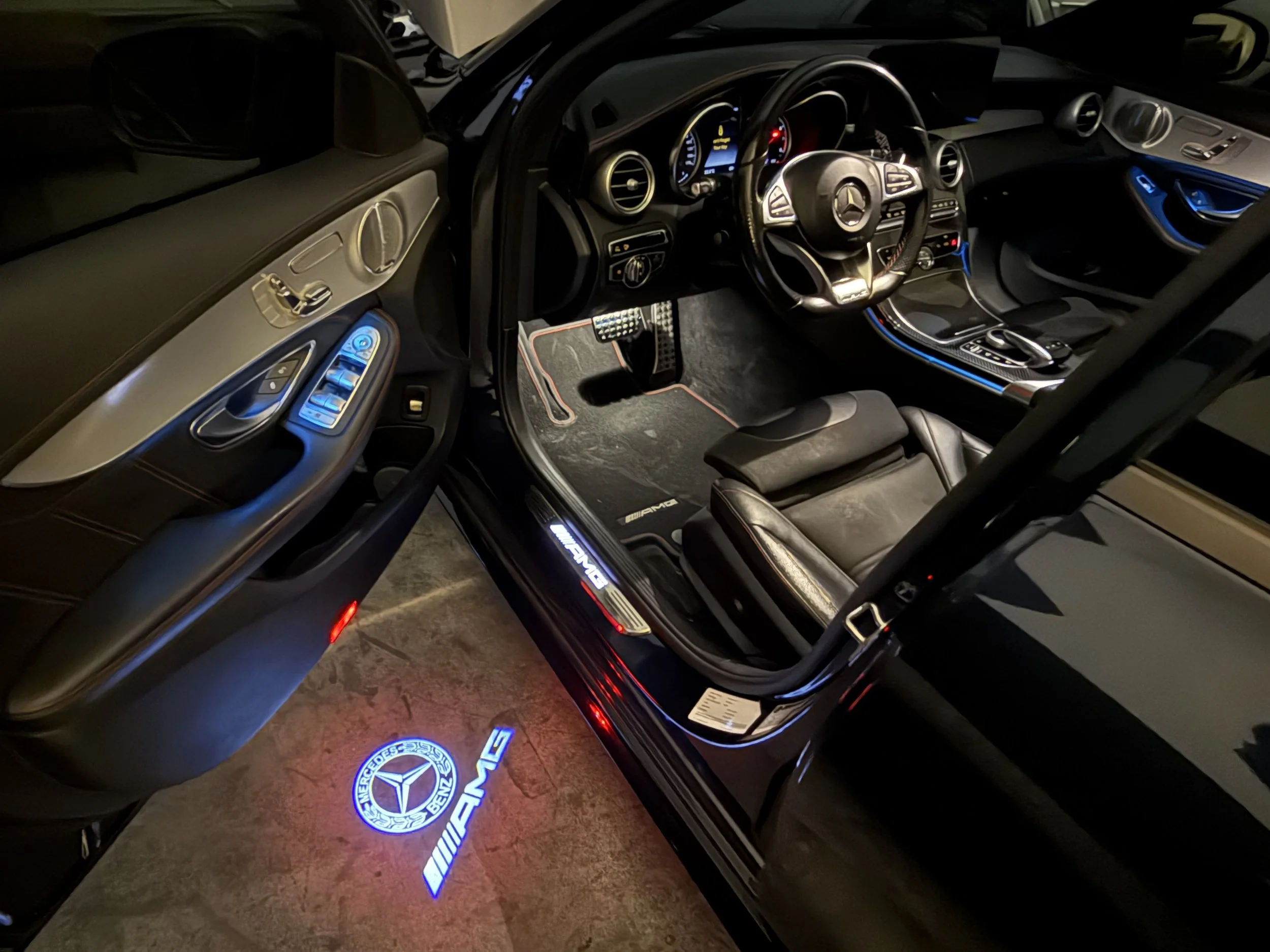 The interior of a Mercedes-Benz AMG car showing the driver seat, steering wheel, dashboard with digital displays, illuminated door controls, and AMG logo projection on the floor with ambient blue lighting.