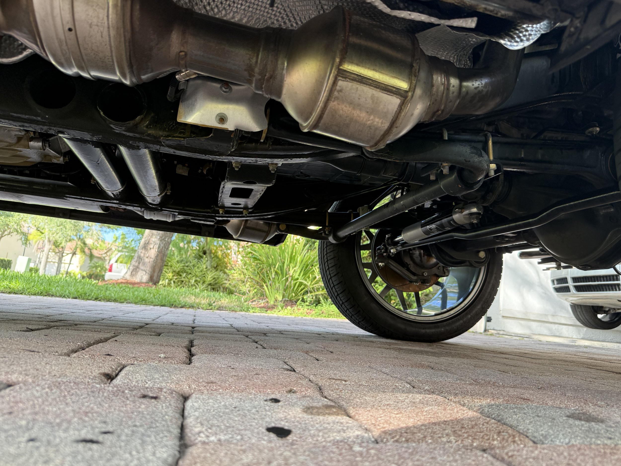 Underneath view of a car showing the exhaust system, suspension, and wheel on a brick driveway with grass and trees in the background.