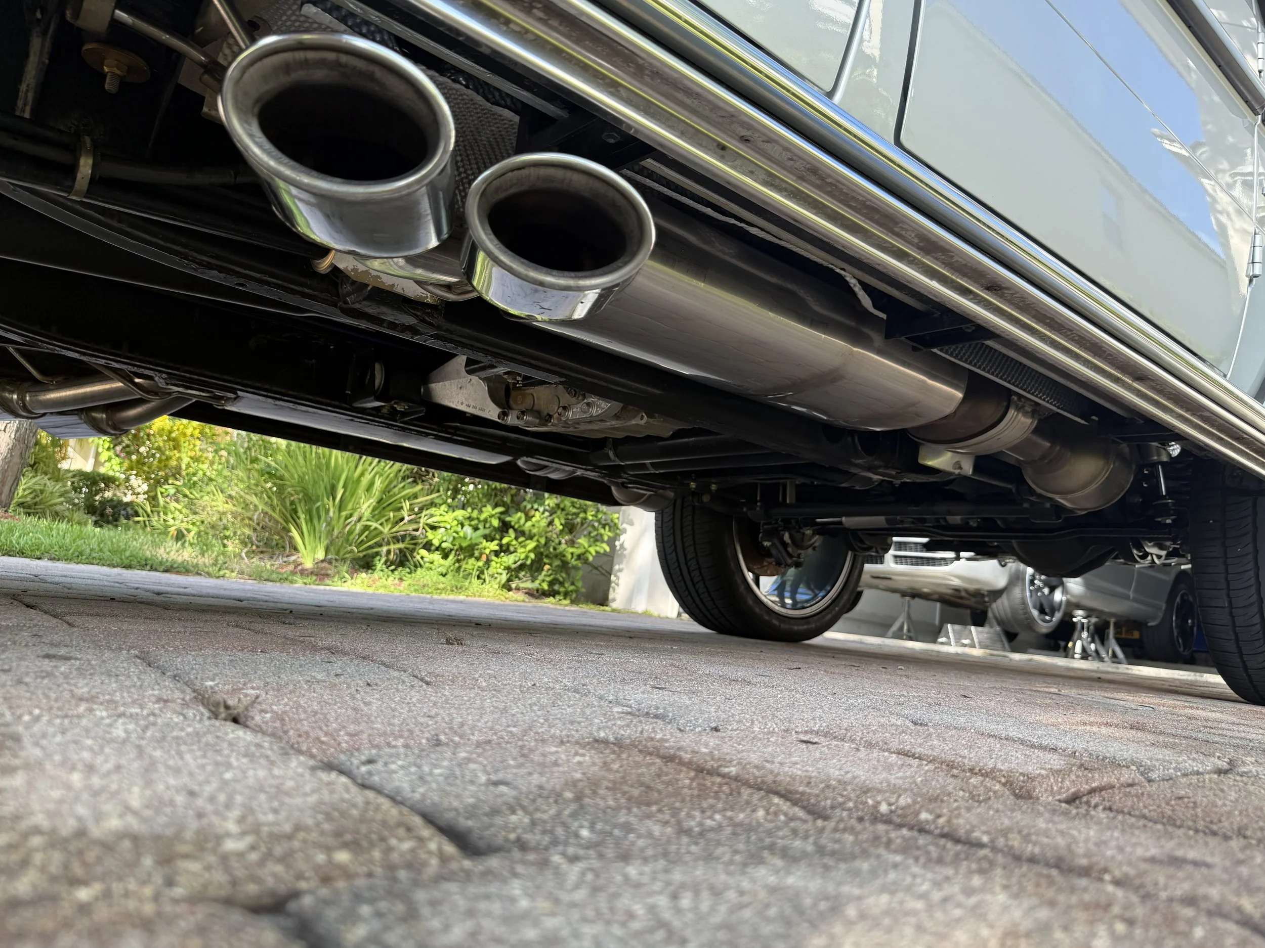 Underneath view of a car focusing on the dual exhaust pipes, with a brick driveway, green plants, and another car visible in the background.