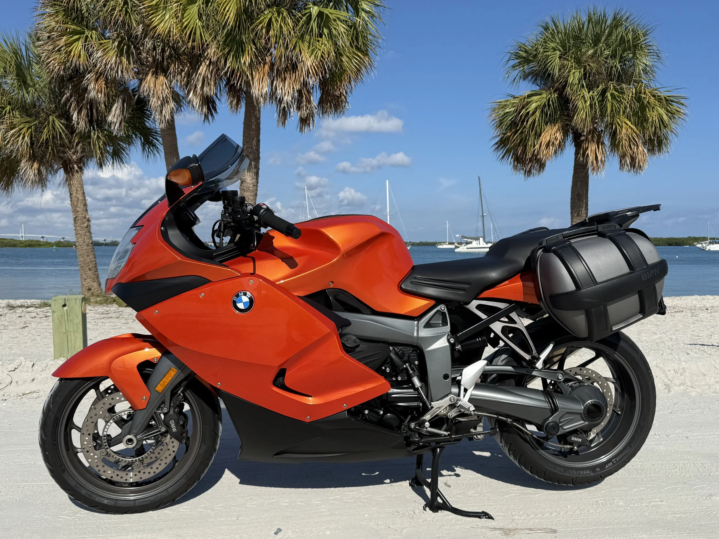 Orange BMW motorcycle parked on a sandy beach with palm trees, a body of water, sailboats, and a blue sky with clouds in the background.