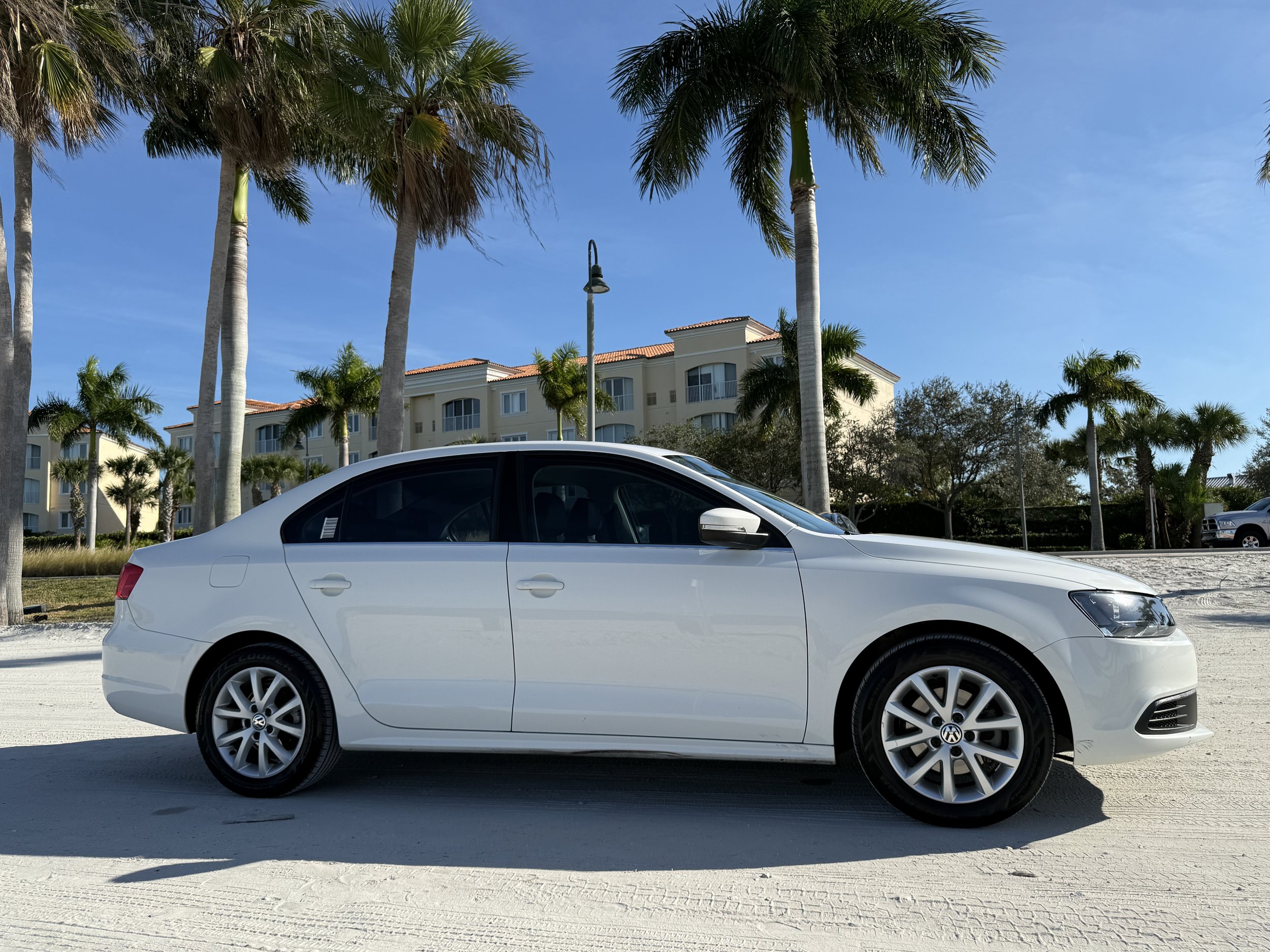 White Volkswagen sedan parked on a sandy surface with palm trees and residential buildings in the background.