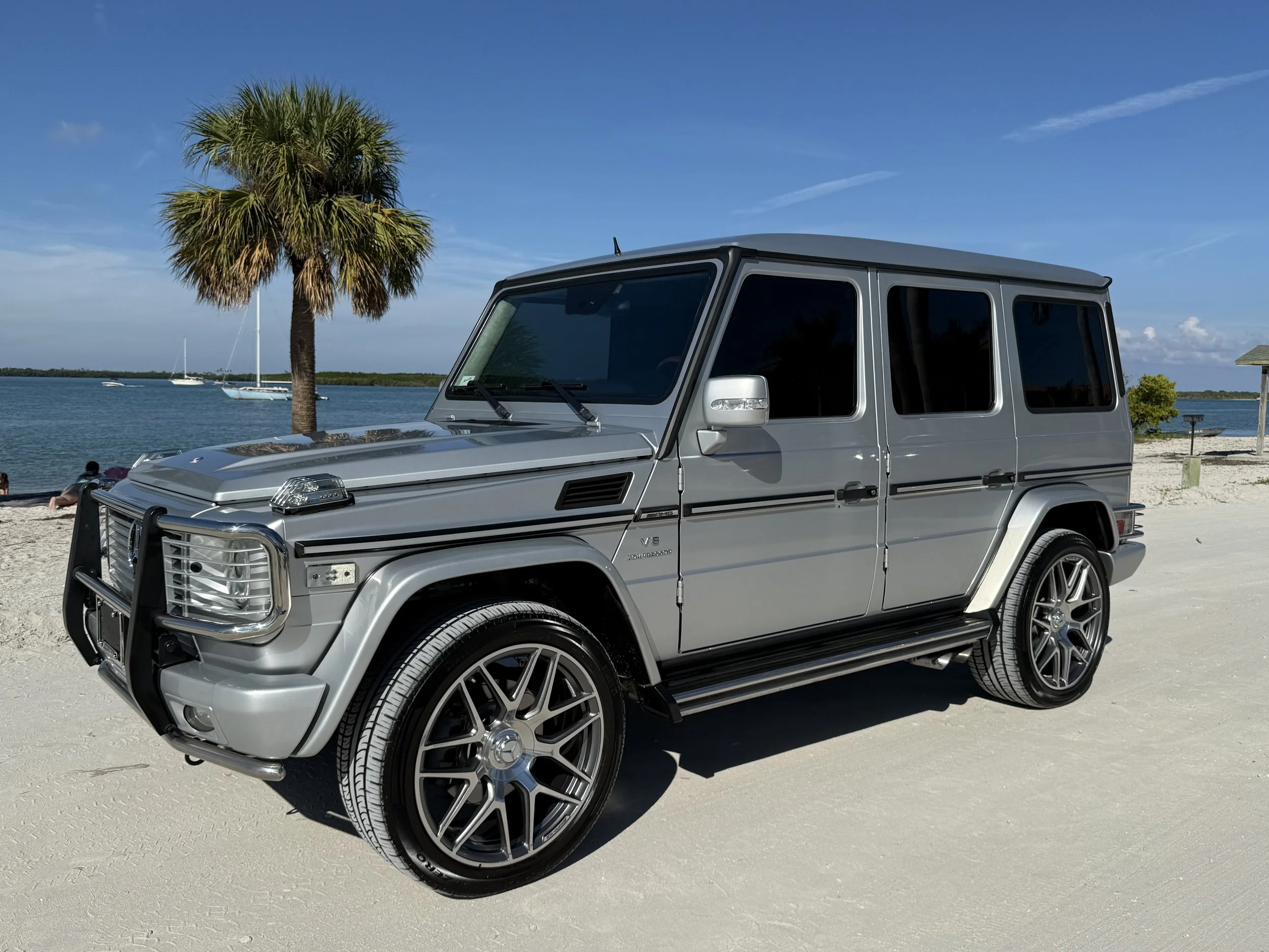 A silver Mercedes-Benz G-Class SUV parked on a sandy beach near a palm tree with water and sailboats in the background.