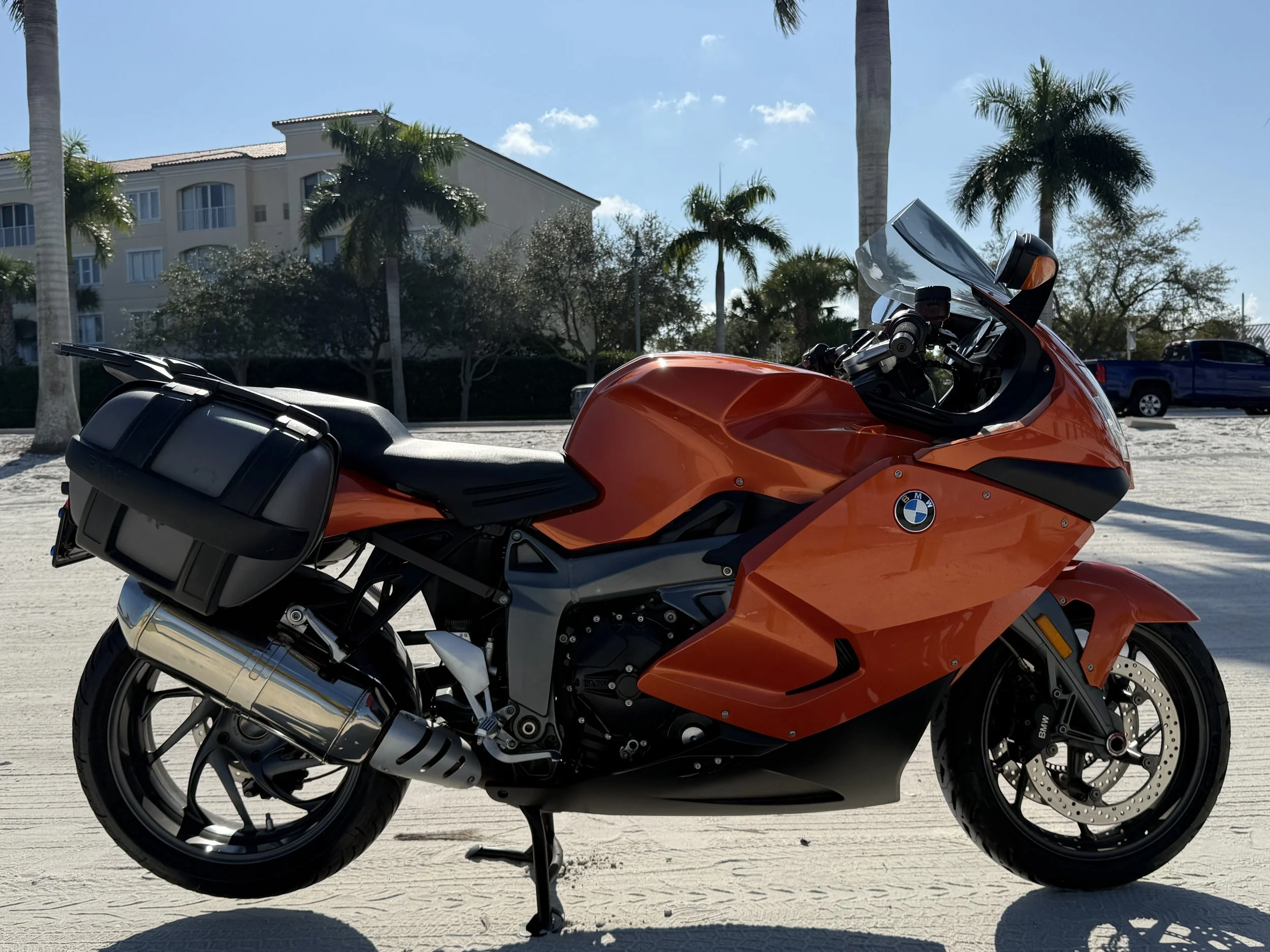 Orange BMW sport motorcycle with black side panniers parked on sandy surface, palm trees, buildings, and a blue truck in the background.
