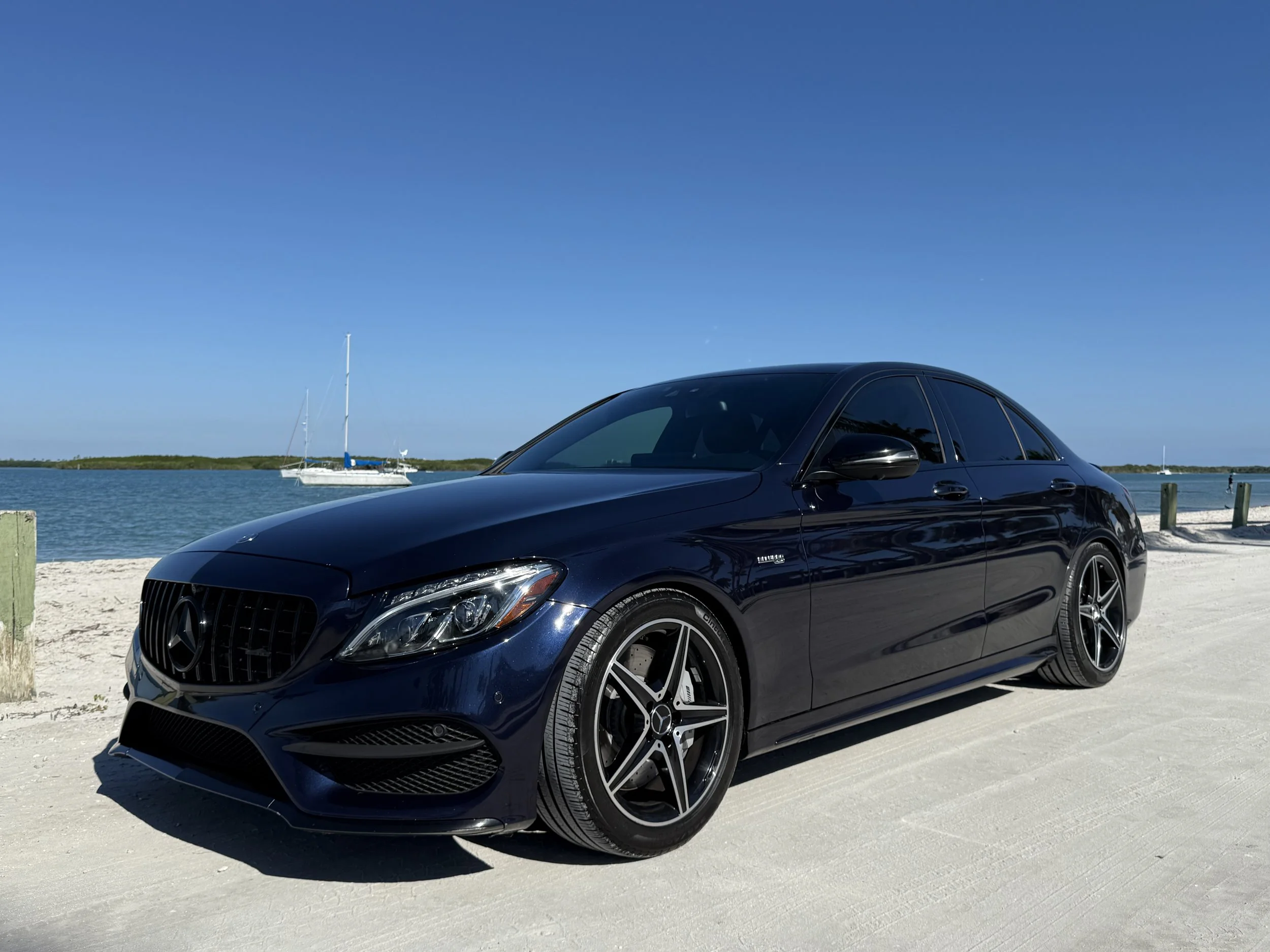 Black Mercedes-Benz sedan parked on sandy beach near water with sailboat in the background under a clear blue sky.