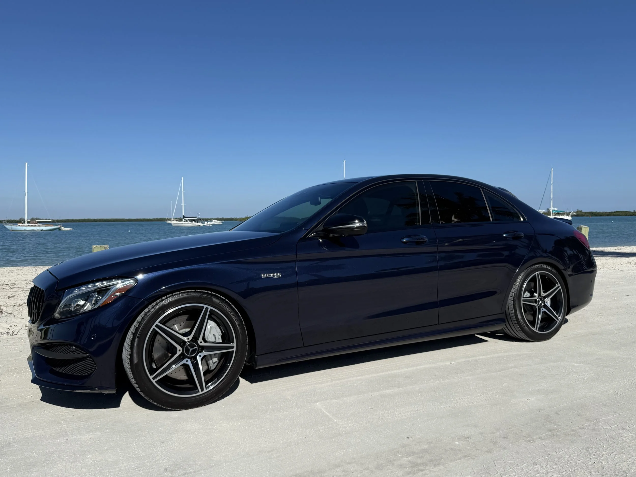 A black Mercedes-Benz sedan parked on a sandy beach with sailboats floating on water and a clear blue sky in the background.