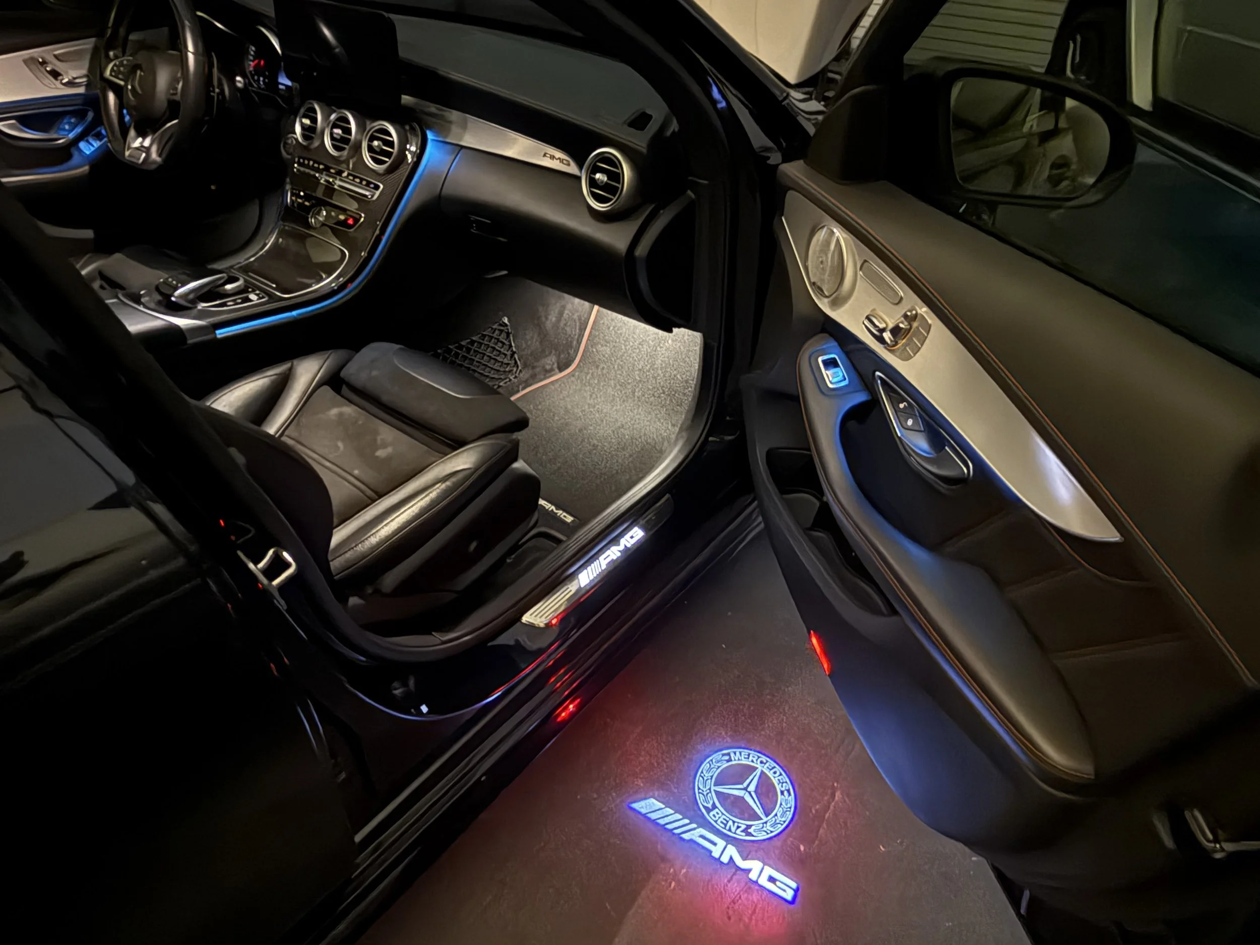 Interior of a black Mercedes-Benz AMG vehicle, showing the dashboard, steering wheel, driver seat, door panel, illuminated AMG door sill, and AMG floor mat with illuminated Mercedes-Benz logo.