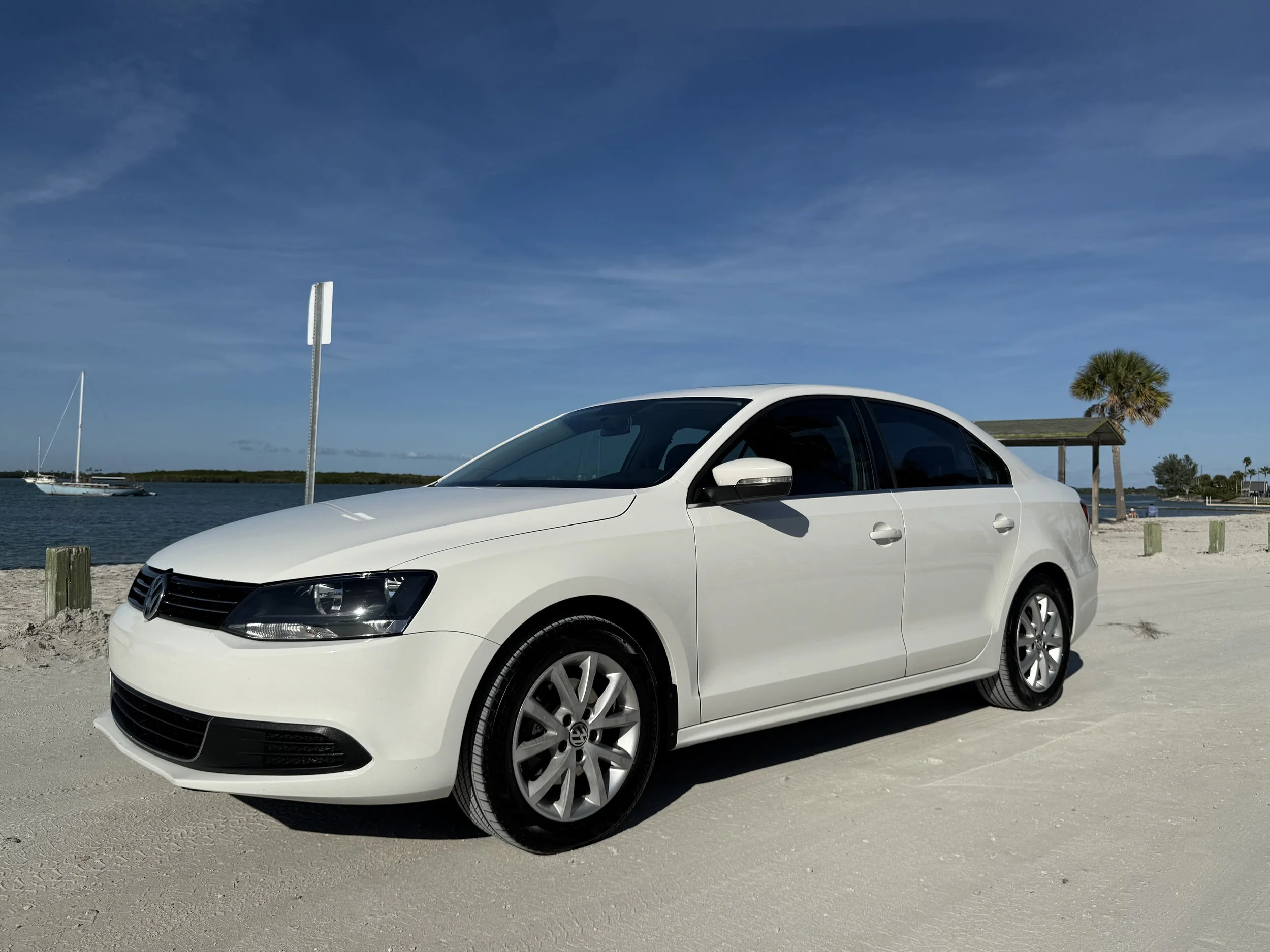 A white Volkswagen sedan parked on sandy beach near water with sailboat, palm trees, and beach shelter in the background under a clear blue sky.