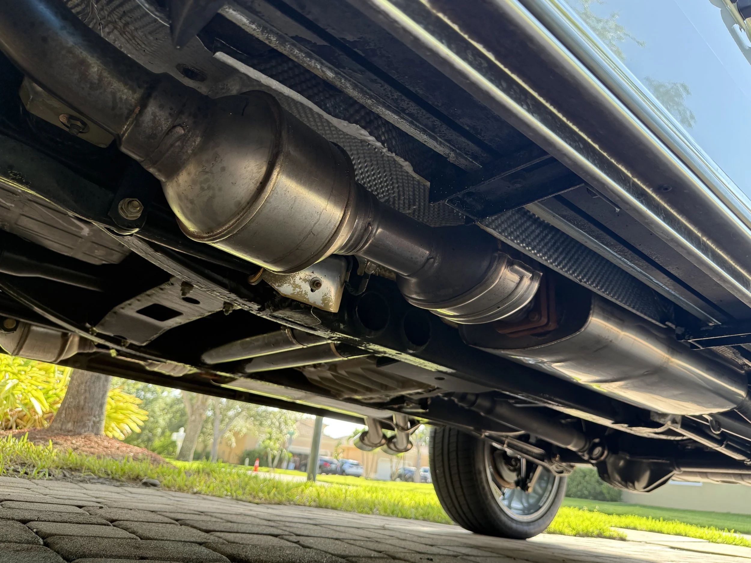 Underneath view of a car showing the exhaust system, muffler, and parts of the suspension on a paved driveway with grass and trees in the background.