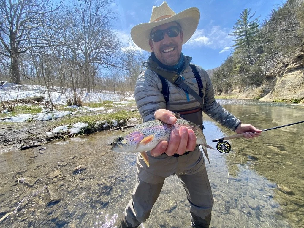 Man wearing a wide-brimmed hat, sunglasses, and outdoor gear smiling and holding a rainbow trout caught while fishing in a river, with trees and a rocky hillside in the background on a sunny day.