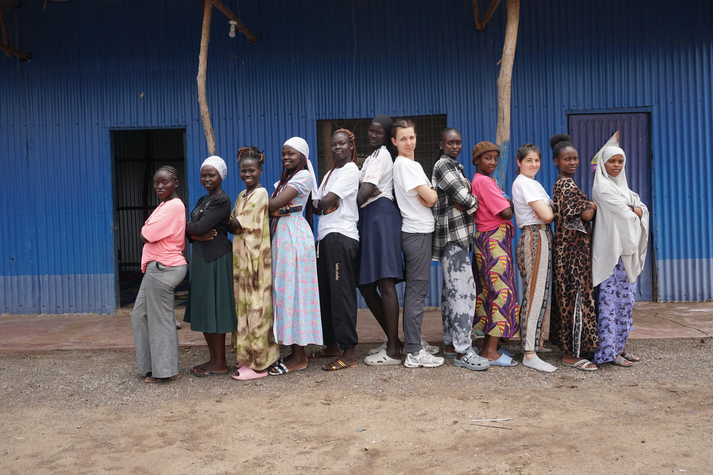 A group of women at an indoor rock climbing gym listening to a woman in black explain something. The gym has climbing walls with holds in bright colors. The women stand on padded mats.