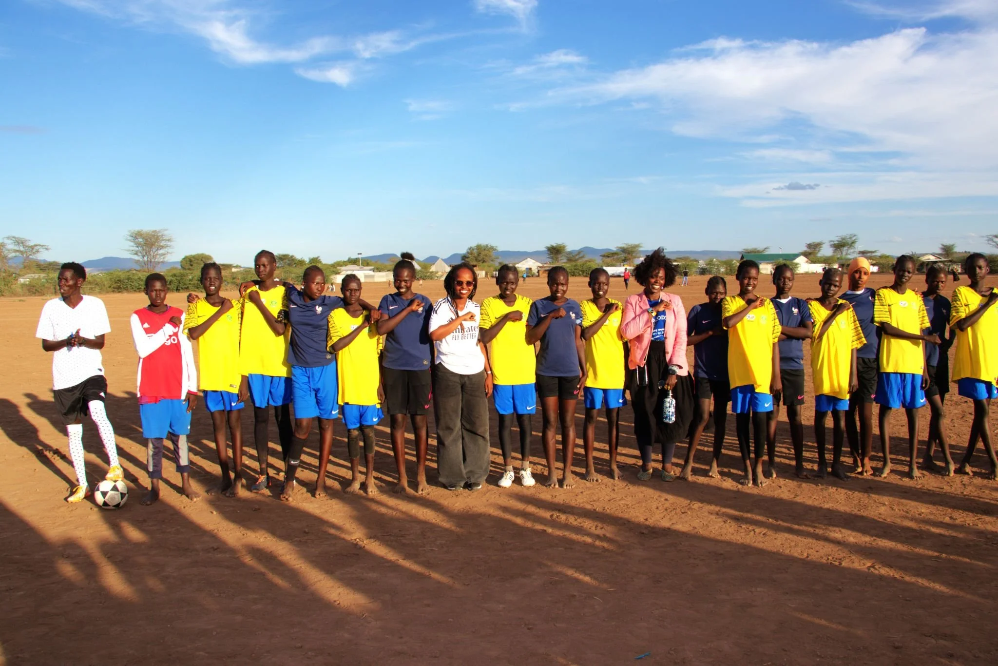 A group of women at an indoor rock climbing gym listening to a woman in black explain something. The gym has climbing walls with holds in bright colors. The women stand on padded mats.