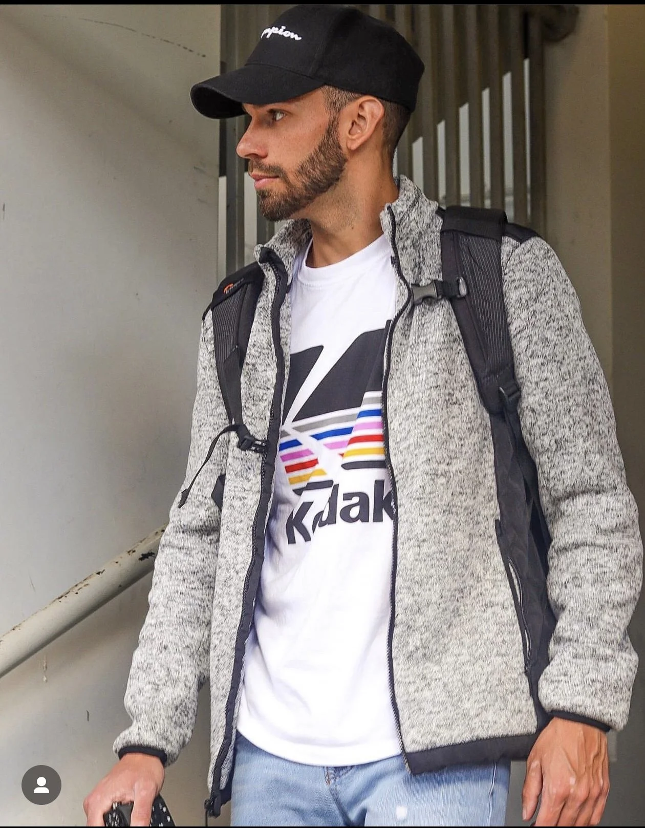 A man wearing a black baseball cap, gray zip-up jacket, white graphic t-shirt, and light blue jeans, carrying a black backpack, standing near stairs.