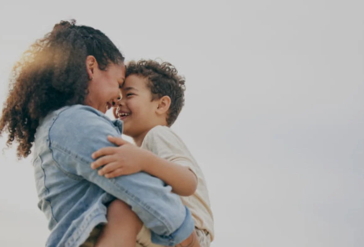 A woman and a young boy are smiling and hugging each other outdoors against a plain, light background.