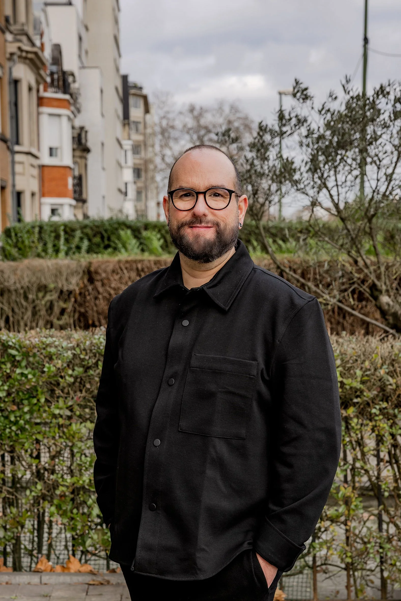 Un homme souriant avec une barbe, portant des lunettes noires et un polo bleu marine, se tient devant un arbuste vert dense.