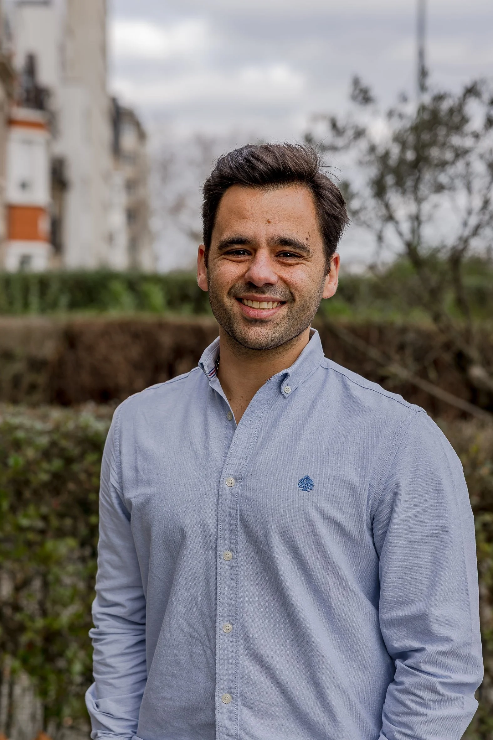 Un homme souriant avec une chemise à rayures blanches et bleues, posant devant un buisson vert.