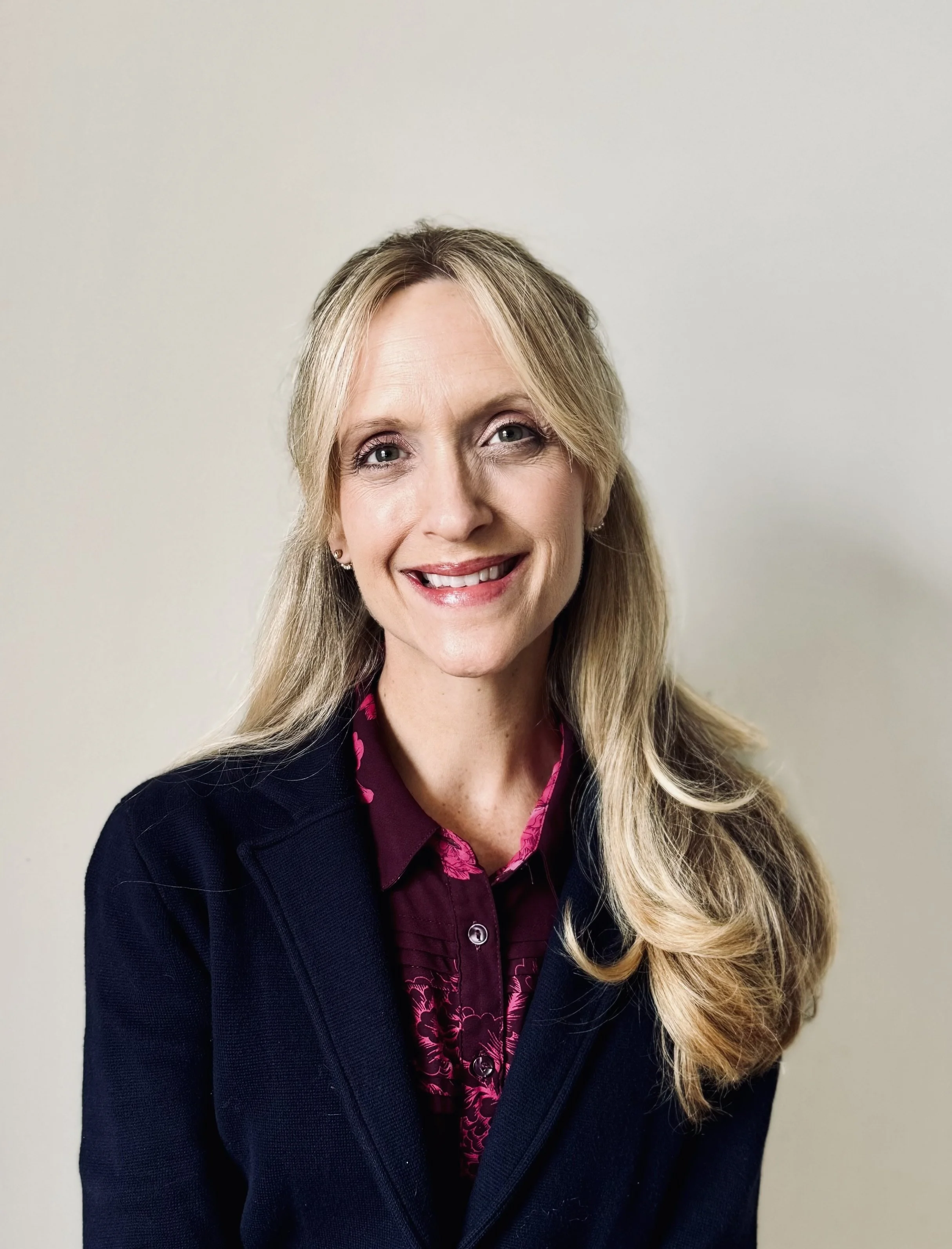 Portrait of a smiling woman with blonde hair, wearing a dark blazer and a purple shirt with pink pattern, standing against a plain beige background.