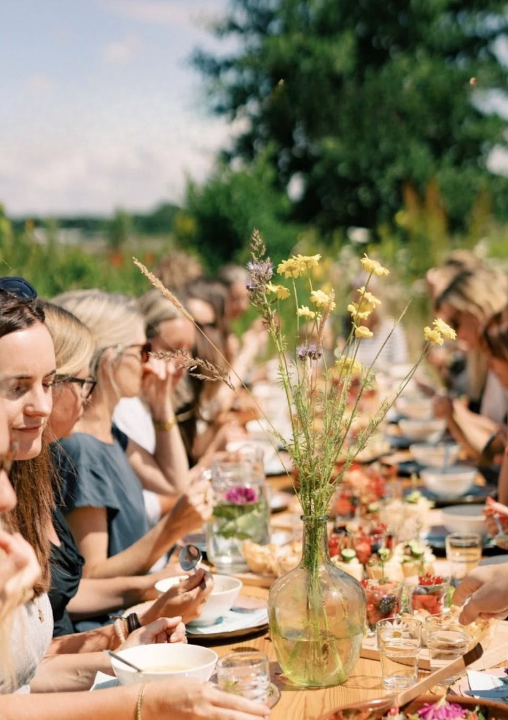 Mensen zitten buiten aan een lange tafel met bloemen en eten.