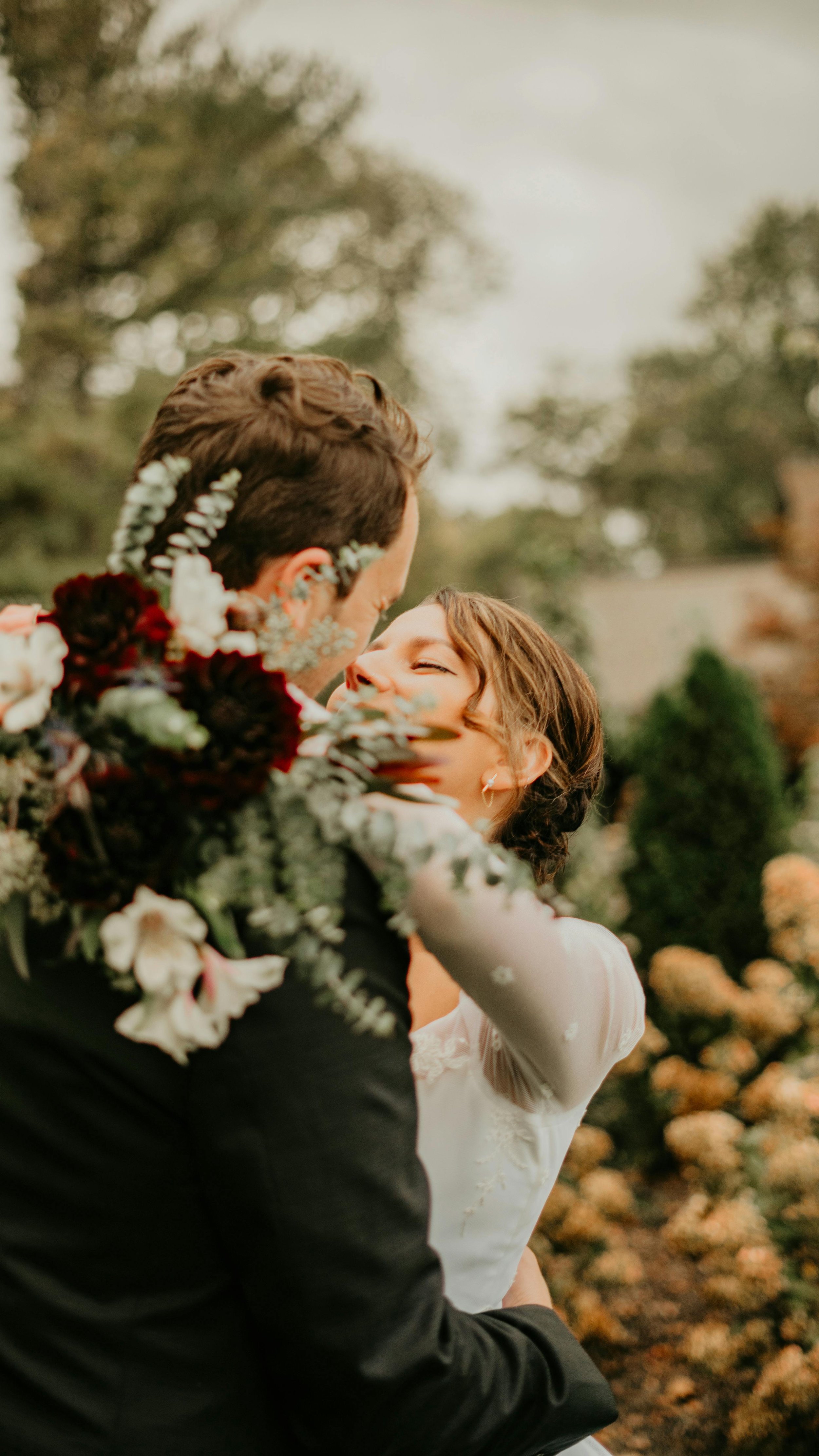 Een stel op een bruiloft omarmt elkaar in een tuin met bloemen. Ze kijken liefdevol naar elkaar, draperie en formele kleding, omgeven door herfstachtige bomen.