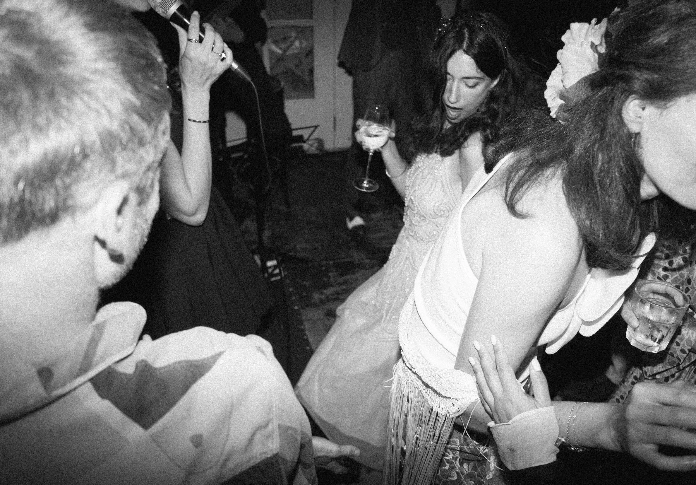 Een groep mensen dansen en genieten op een feestje. Eén vrouw houdt een glaasje wijn vast, anderen drinken uit glazen, terwijl ze samen dansen.