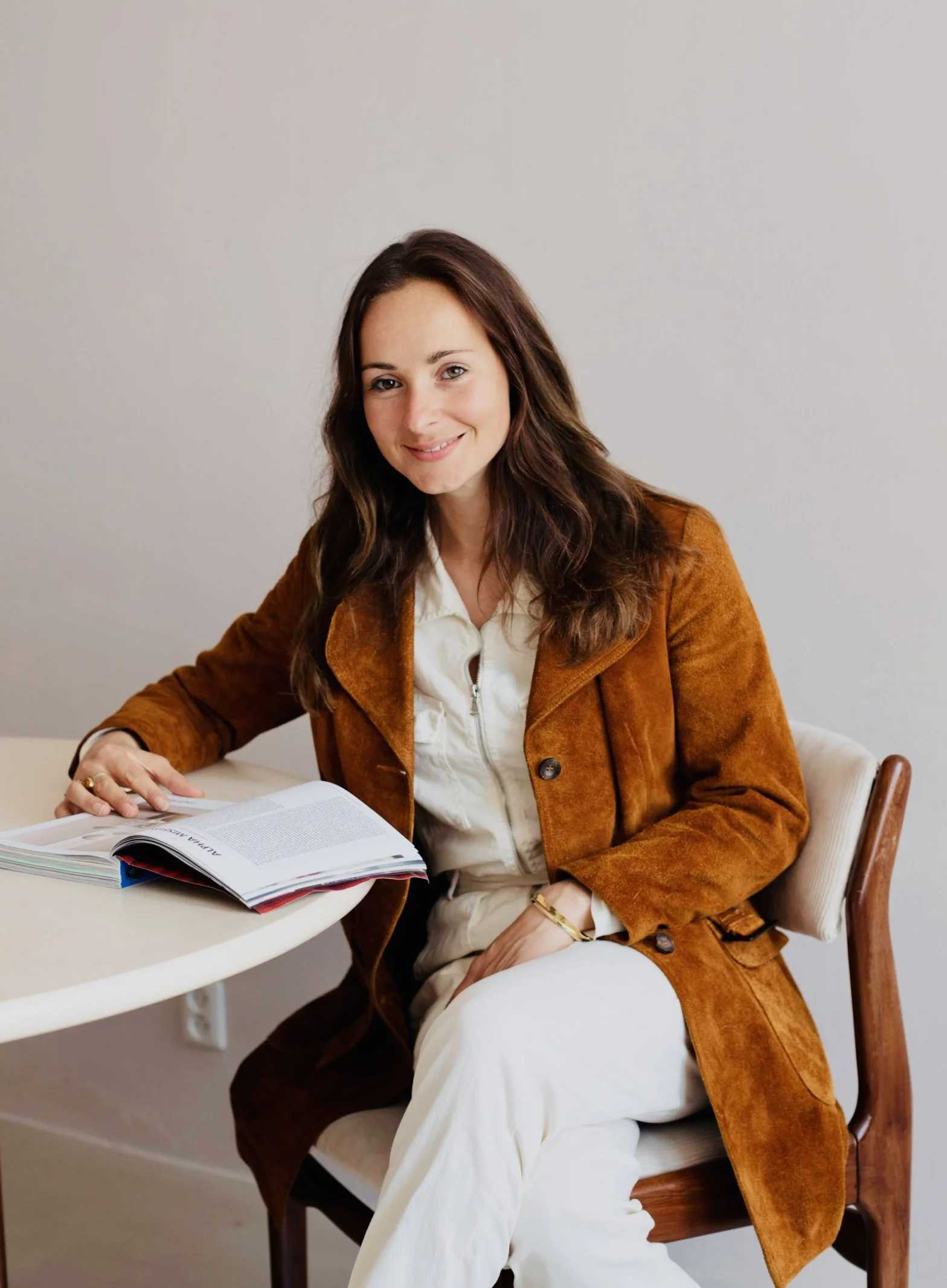 Een vrouw met bruin haar zit aan een ronde tafel met een open boek voor haar, lachend naar de camera, gekleed in een witte blouse en een bruin leren jasje.