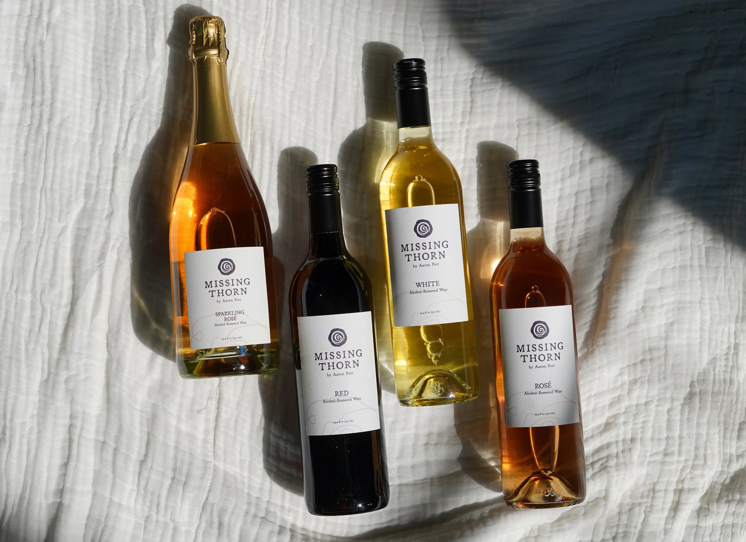 Four bottles of wine with white labels from the brand 'Missing Thorn' placed on a textured white fabric, with sunlight casting shadows.