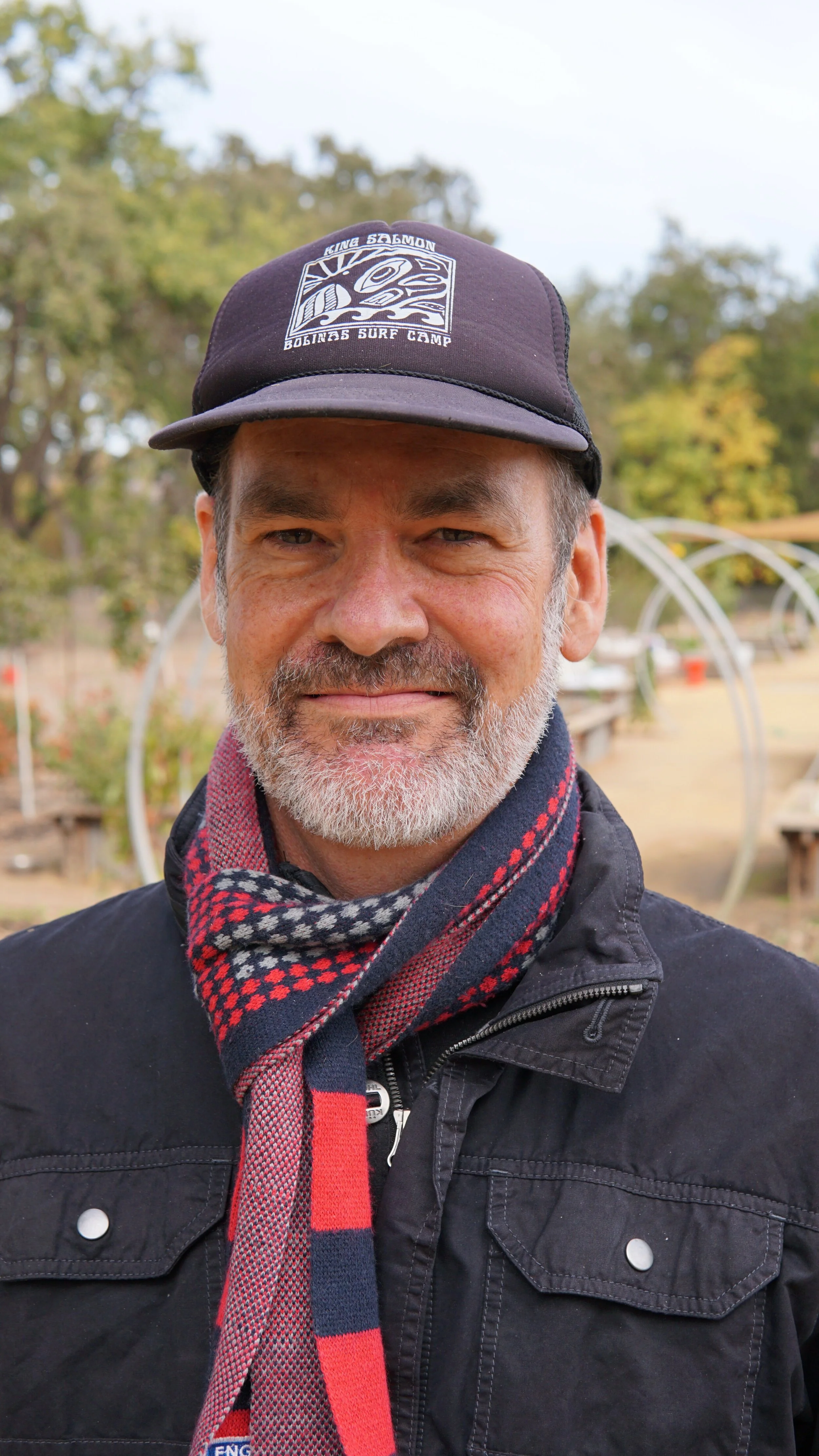 Aaron Pott, wearing a black jacket, a patterned scarf, and a navy cap with a logo that says 'King Salmon Boat & Surf Camp'. He is outdoors in a natural setting with trees and outdoor structures in the background.