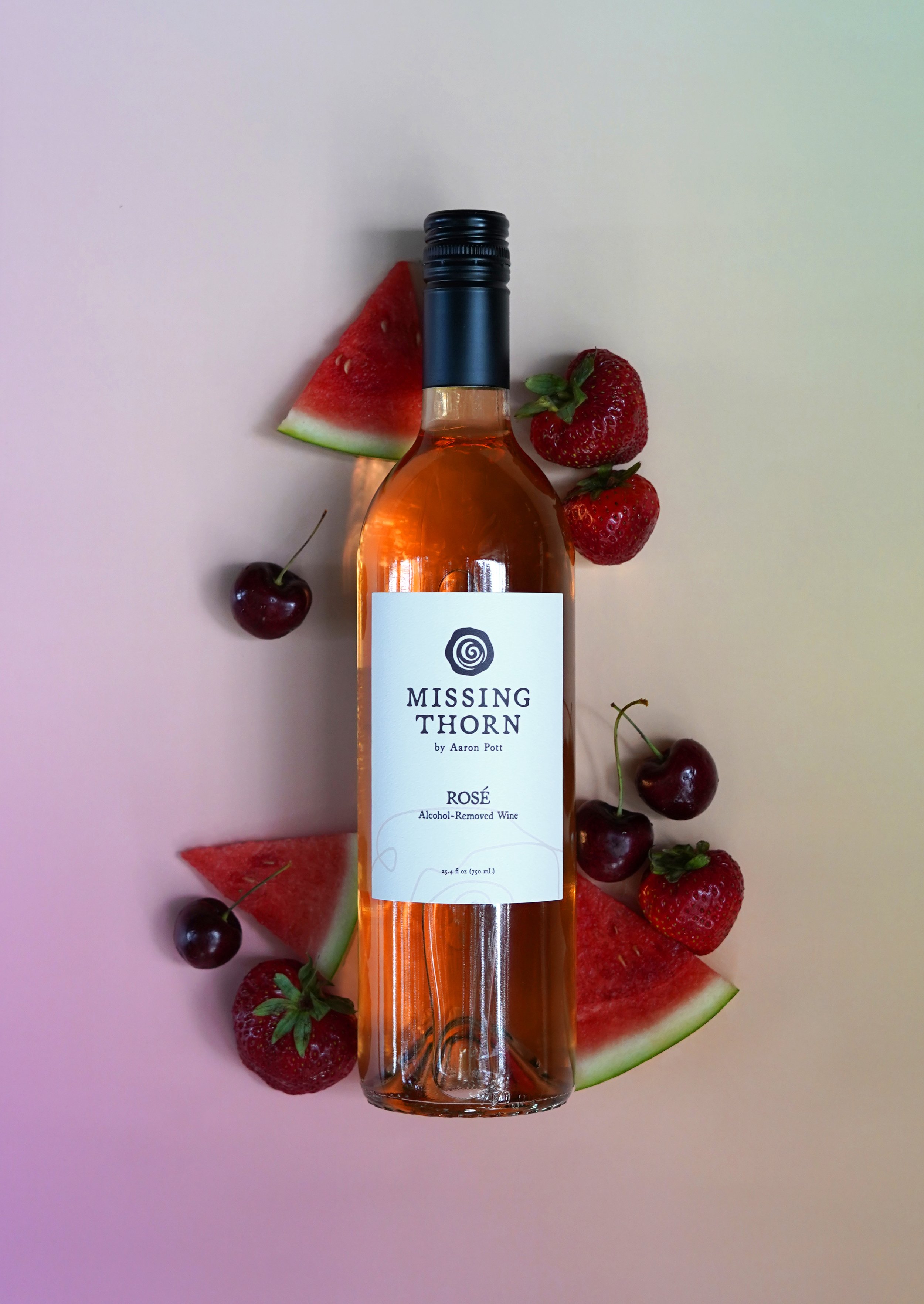 A bottle of Missing Thorn Rosé wine surrounded by slices of watermelon and strawberries, with cherries scattered around on a light-colored surface.