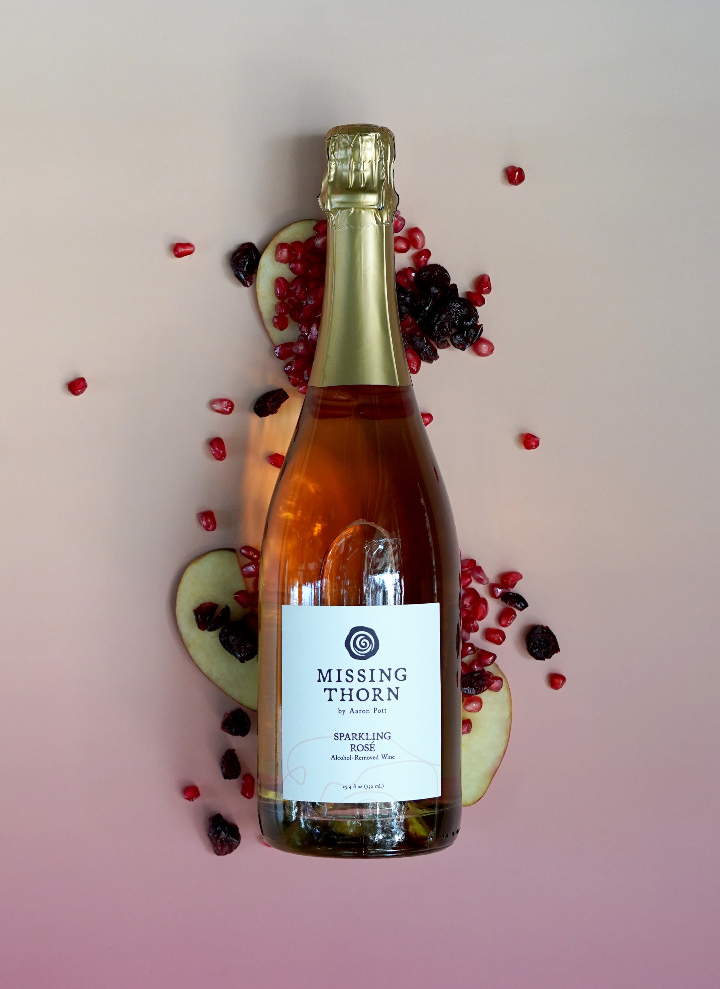 A bottle of sparkling rosé wine labeled "Missing Thorn by Aaron Pott" surrounded by slices of apple, pomegranate seeds, and dried cranberries on a light pink background.