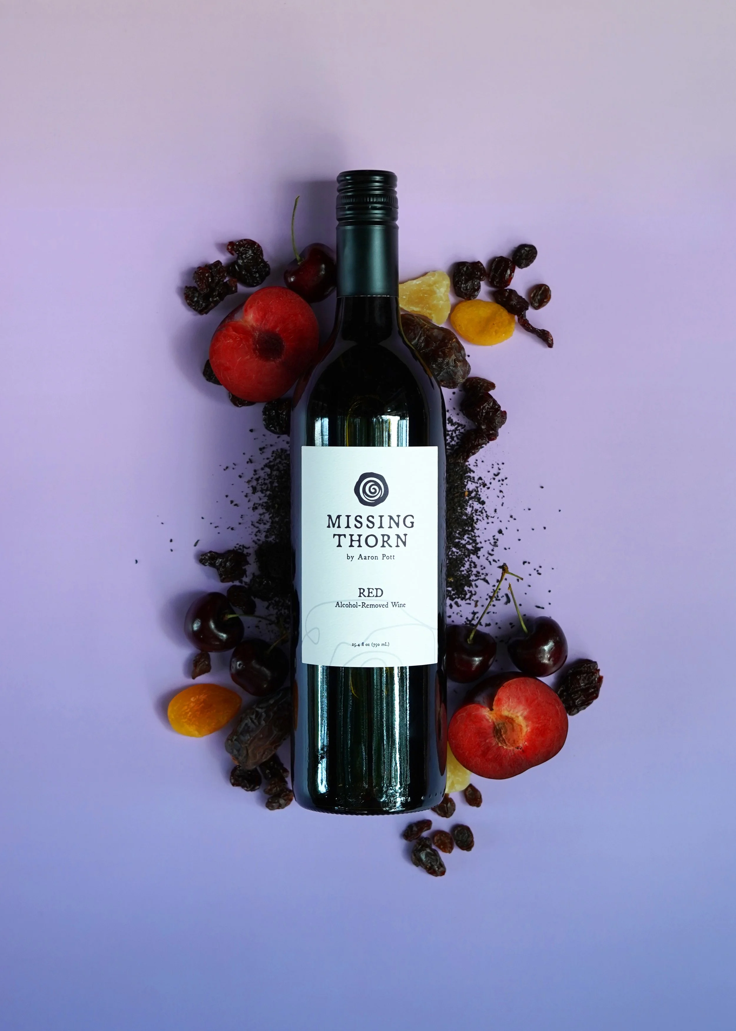 A bottle of Missing Thron red wine surrounded by cherries, apricots, raisins, and crushed black seeds on a purple background.