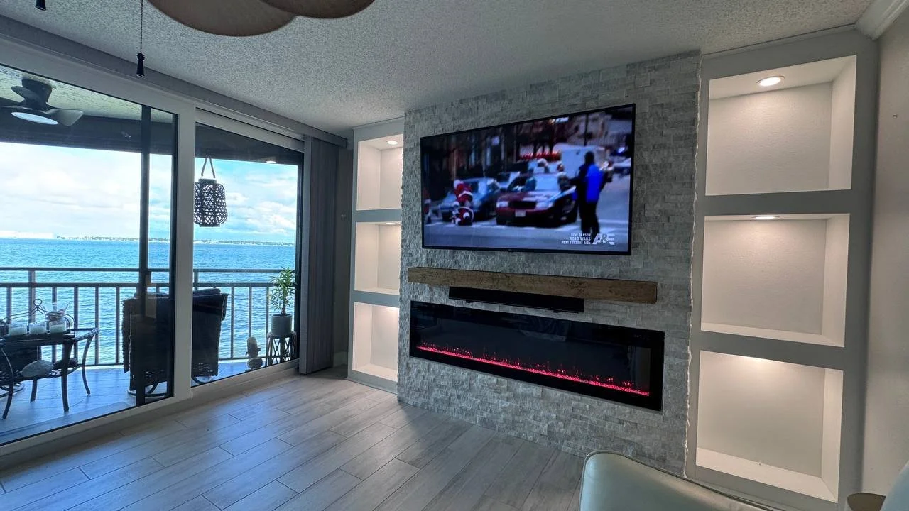 A full-height media wall in Sarasota with central stacked stone, electric fireplace, TV, wood mantel, and built-in display niches with lighting.