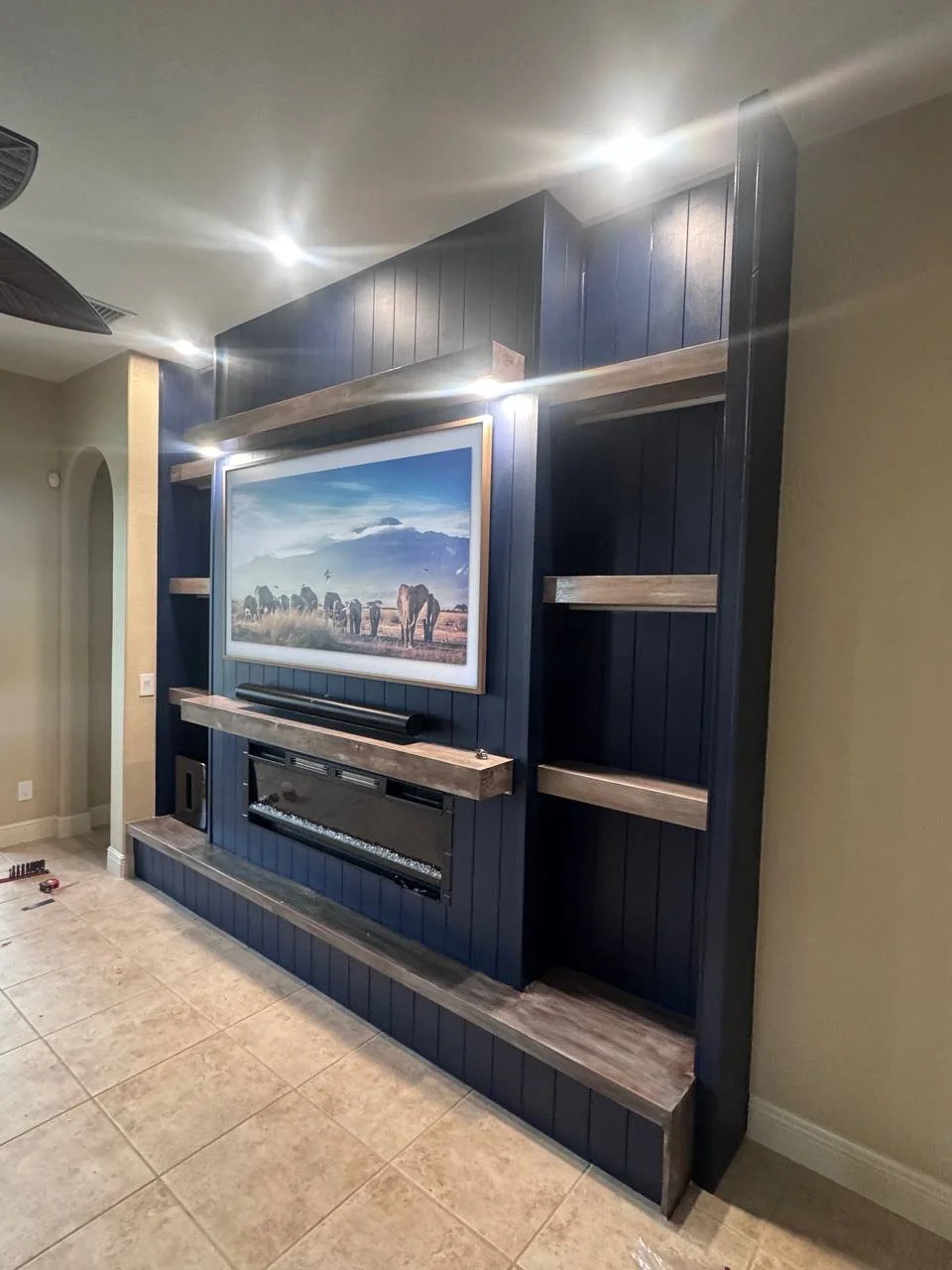 Side perspective of a navy fireplace media wall in Dallas showing illuminated niches, horizontal wood shelves, a frame television, and a linear fireplace.