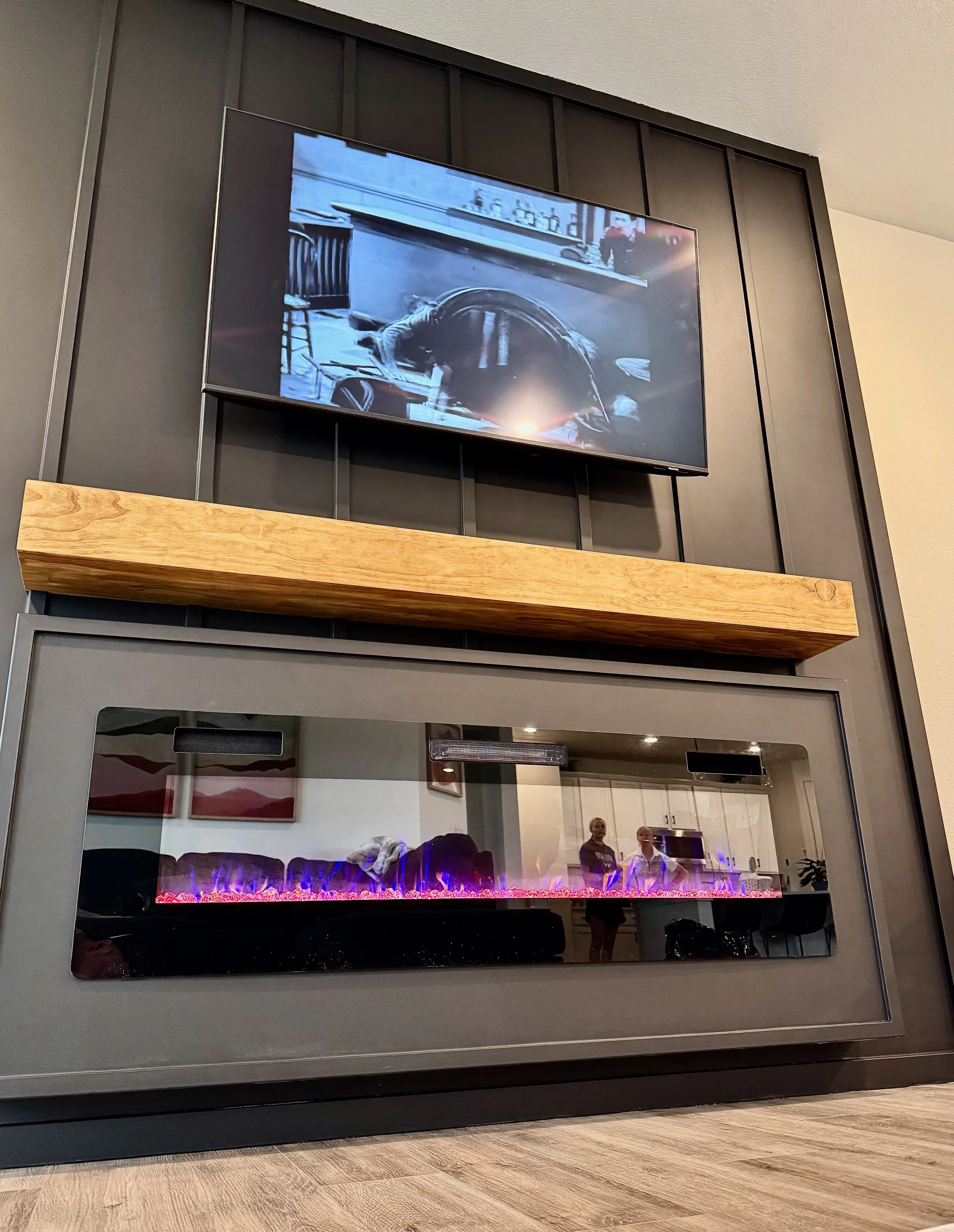 A modern interior fireplace with a digital flame display, a wooden mantle, and a mounted television above it, showing a black and white scene.