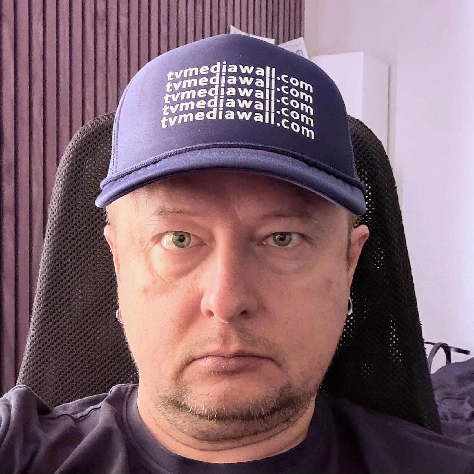 A person wearing a dark blue baseball cap with 'tvmediawall.com' written repeatedly, sitting in a black mesh-backed chair in an indoor setting.