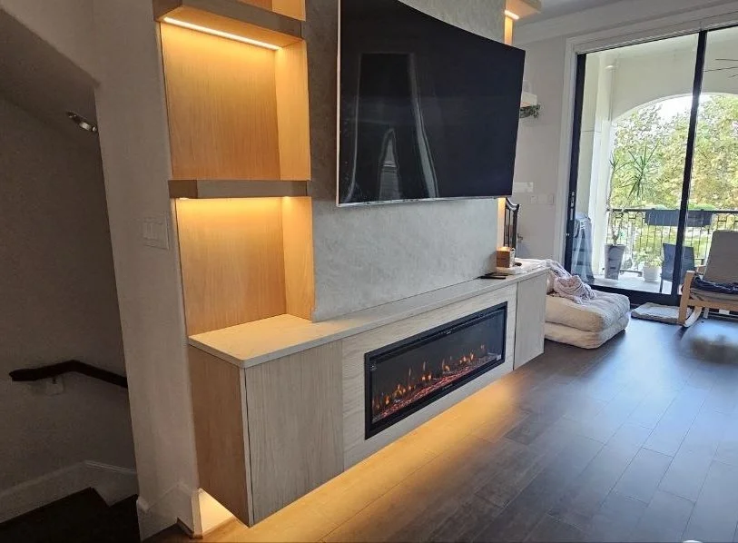 Angled view of a Houston entertainment built-in with an electric fireplace, floating storage cabinet, illuminated wood shelves, and decorative TV panel