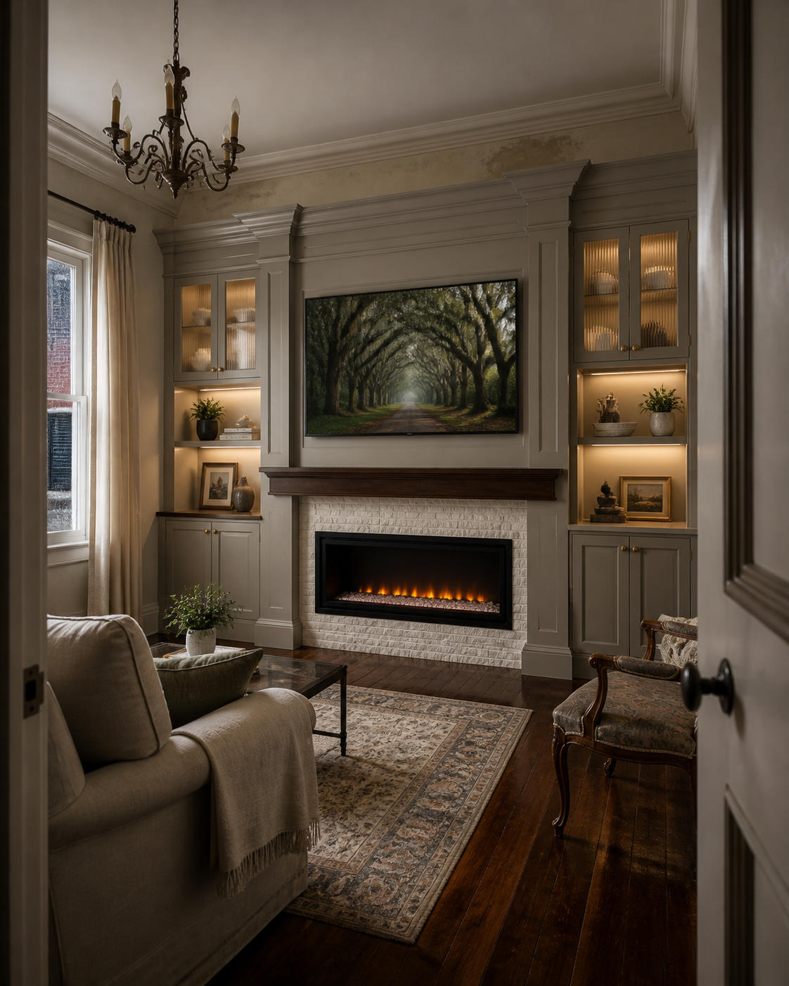 Living room with white brick fireplace, TV displaying an outdoor scene with tree-lined path, built-in shelves with decor, beige sofa with throw blanket, armchair, chandelier, window with curtains, wooden flooring, and rug.