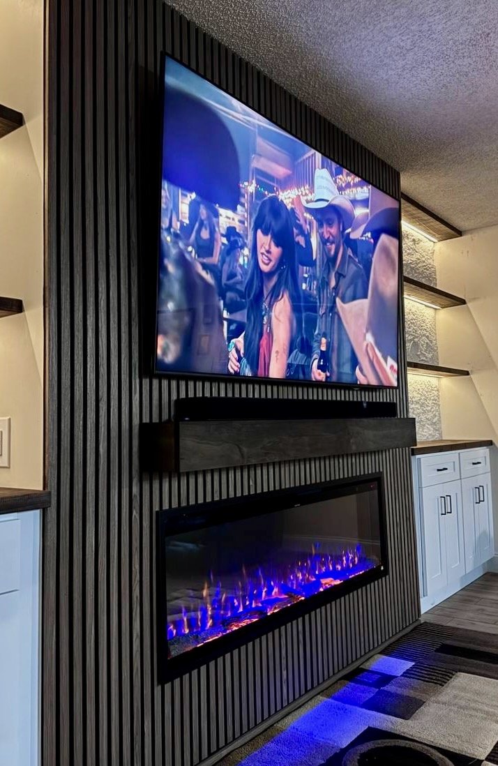 Angled view of a black slat fireplace media wall with a mounted TV, dark wood mantel, electric fireplace, and illuminated built-in shelving in Tampa.